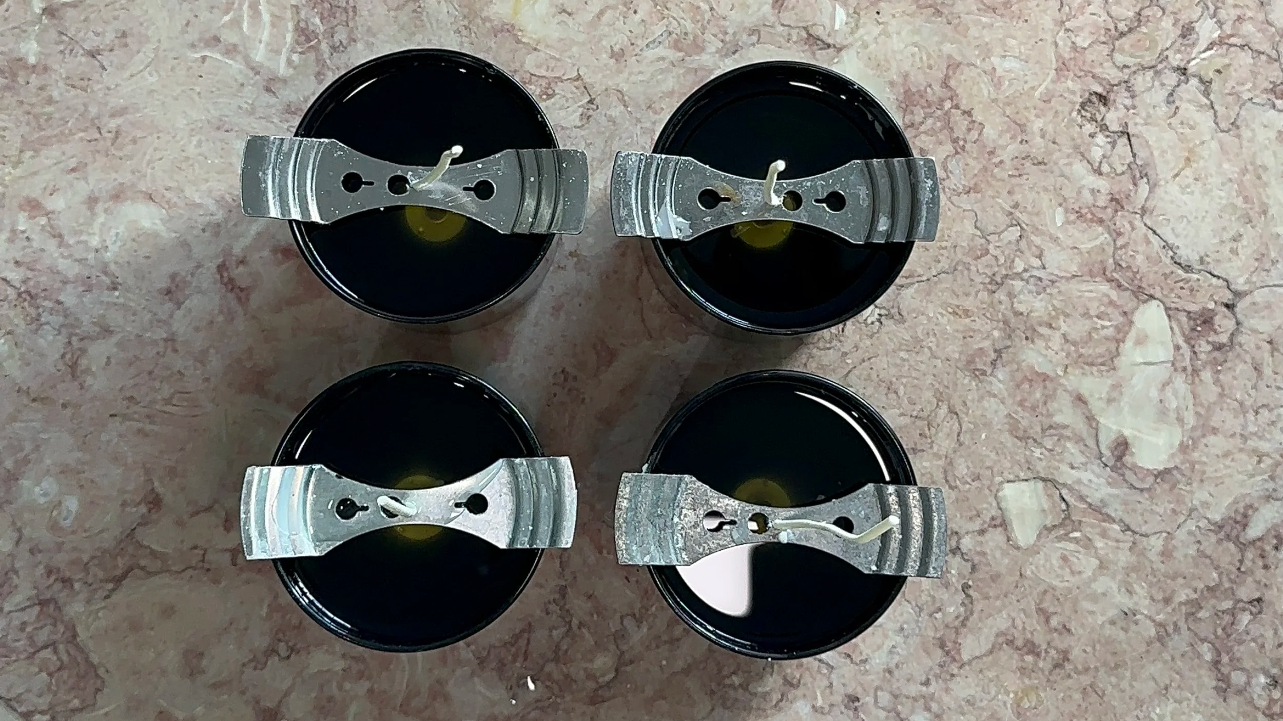 Four black containers filled with dark liquid, each sealed with metallic lids and wicks, arranged on a pinkish marble surface.