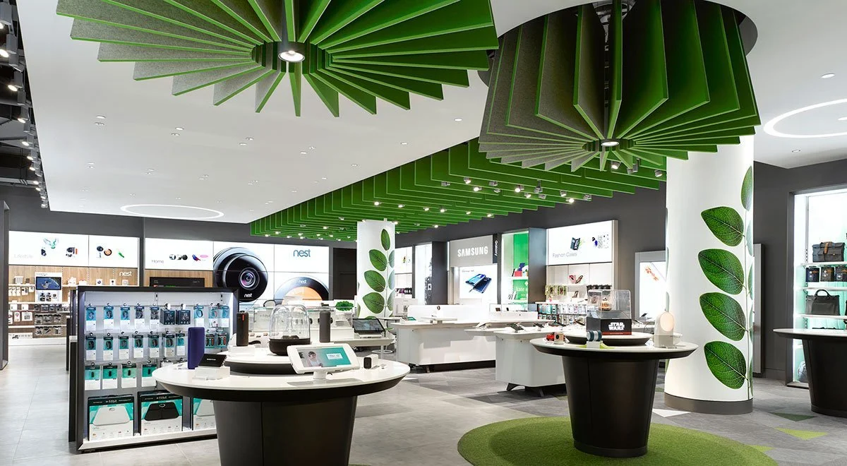 Interior of an electronics retail store with green and white decor, display tables with electronic devices, large screens on the wall, and ceiling installations resembling green leaves.