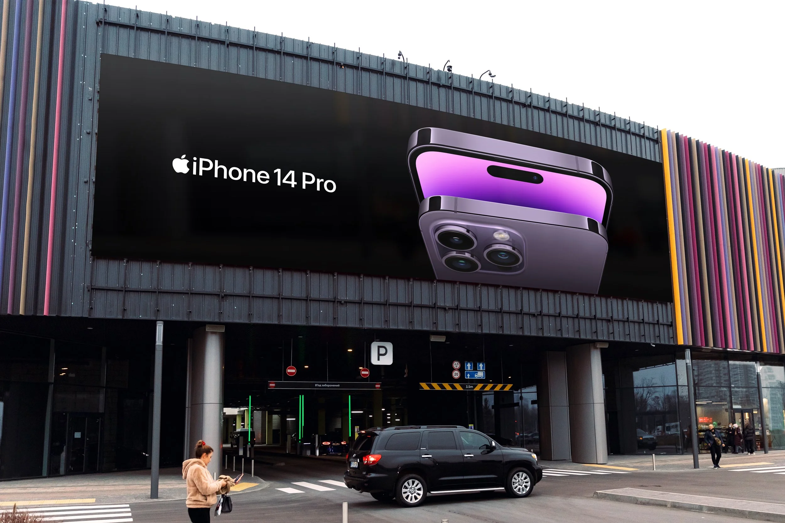 Large advertisement for the iPhone 14 Pro on a building's exterior, showing the phone with a purple and black color scheme. There are pedestrians in the foreground, including a woman with a backpack, and a black SUV parked nearby.