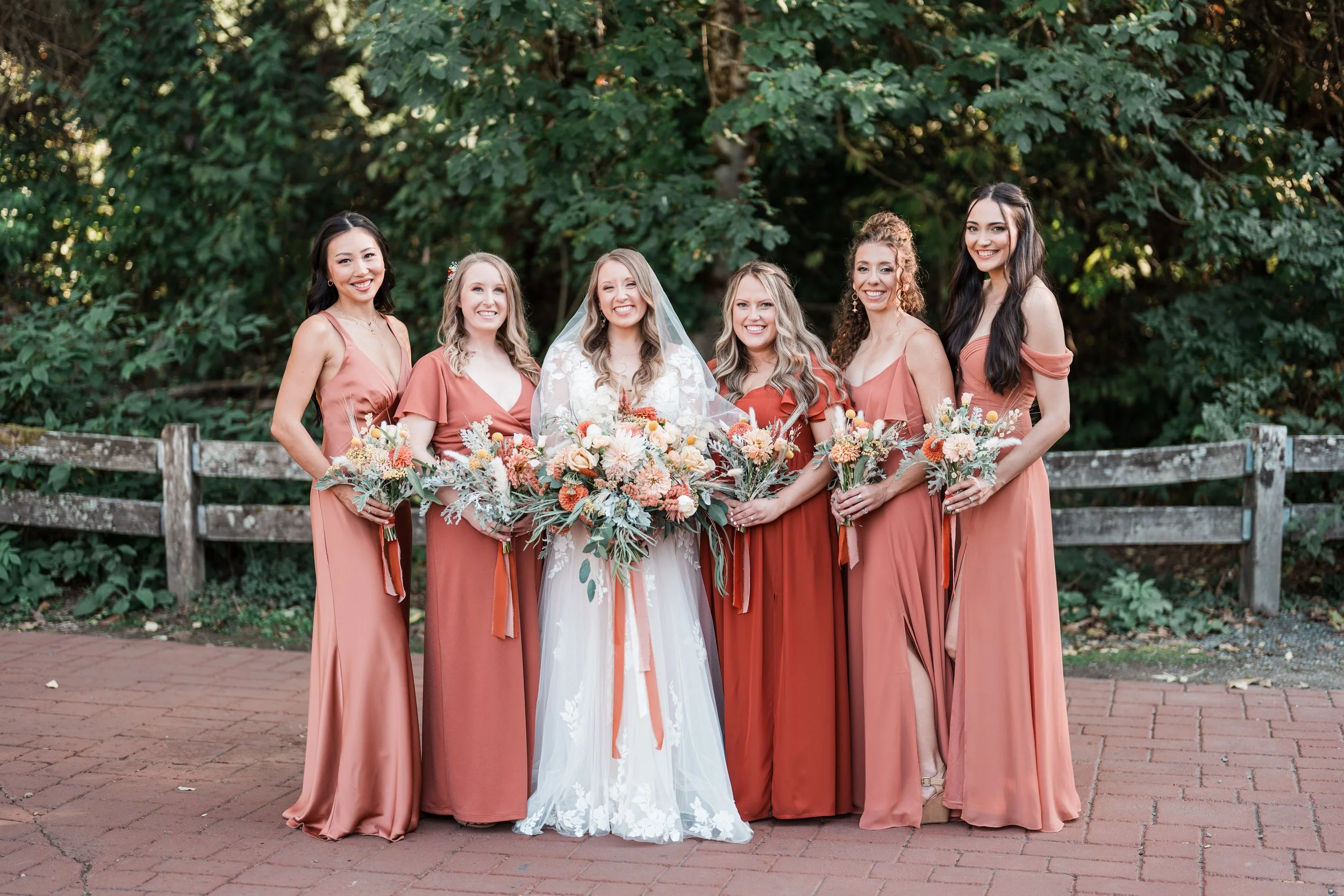 A bride in a white wedding dress holding a bouquet of flowers standing with five bridesmaids in matching terracotta dresses, each holding a bouquet, outdoors with a wooden fence and green foliage background.