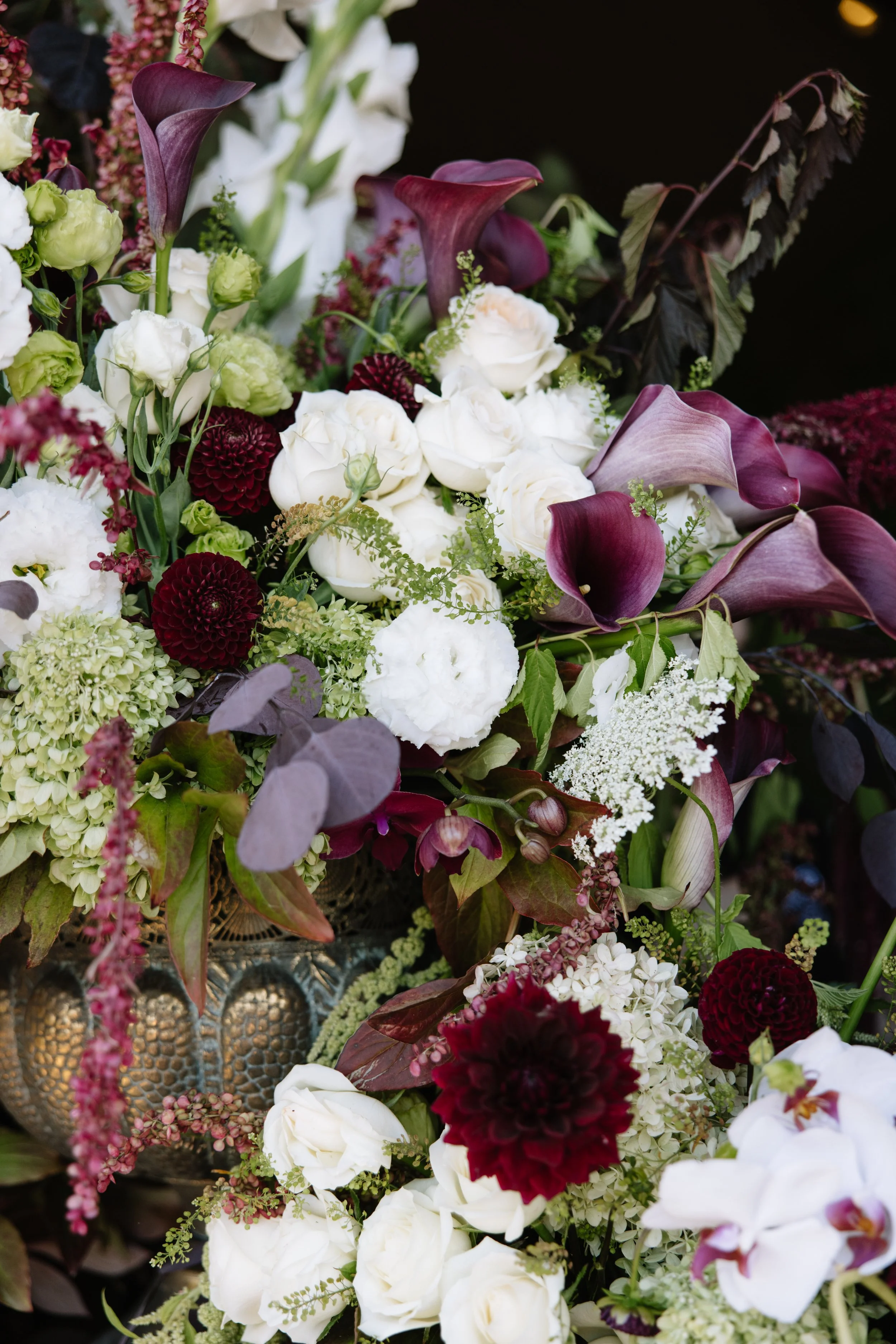 Close-up of a wedding floral arrangement with white roses, dark purple calla lilies, burgundy dahlias, and green foliage in a decorative metallic vase.