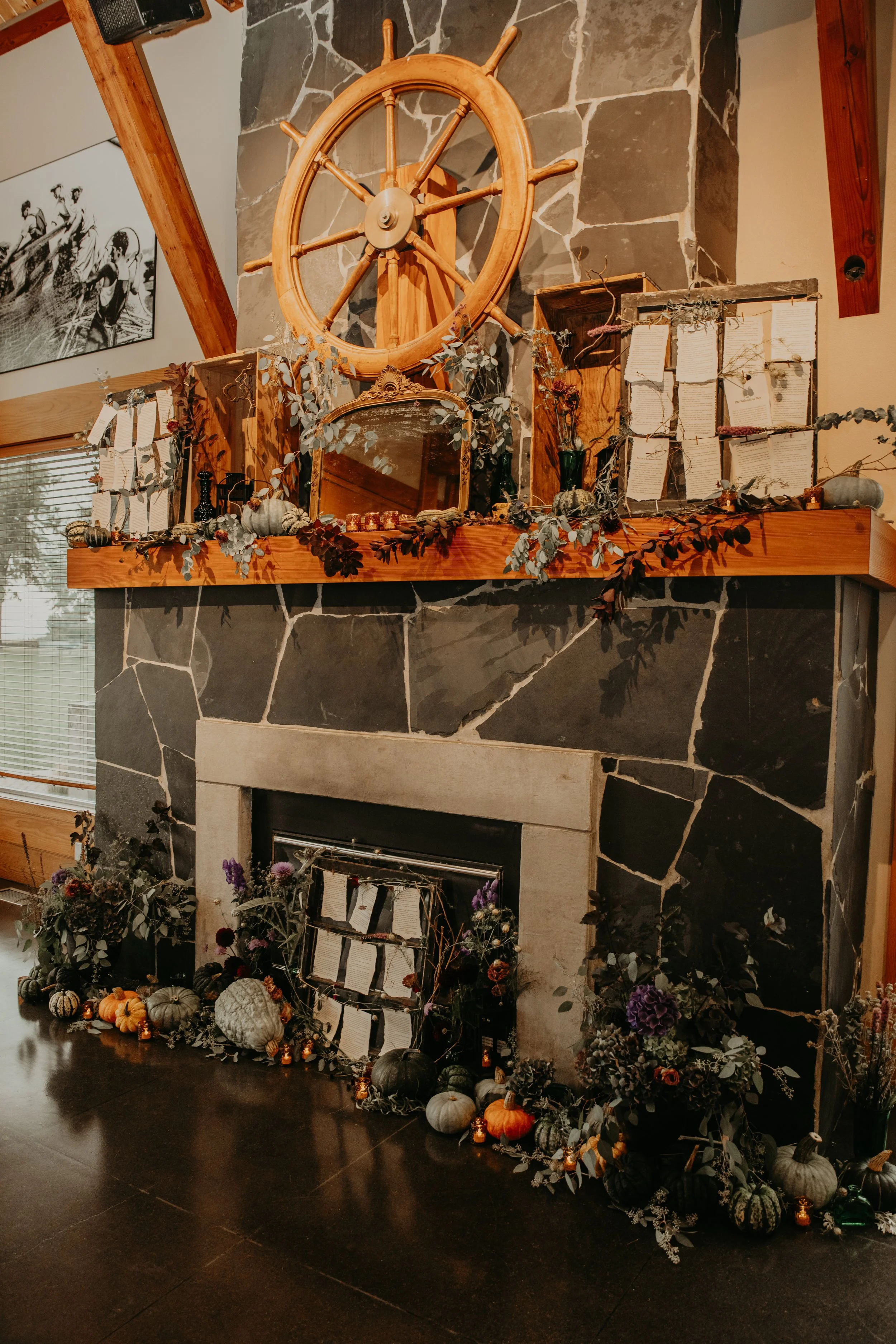 Decorated fireplace for a wedding with pumpkins, plants, and hanging notes or letters, with a nautical ship wheel mounted above the mantel.