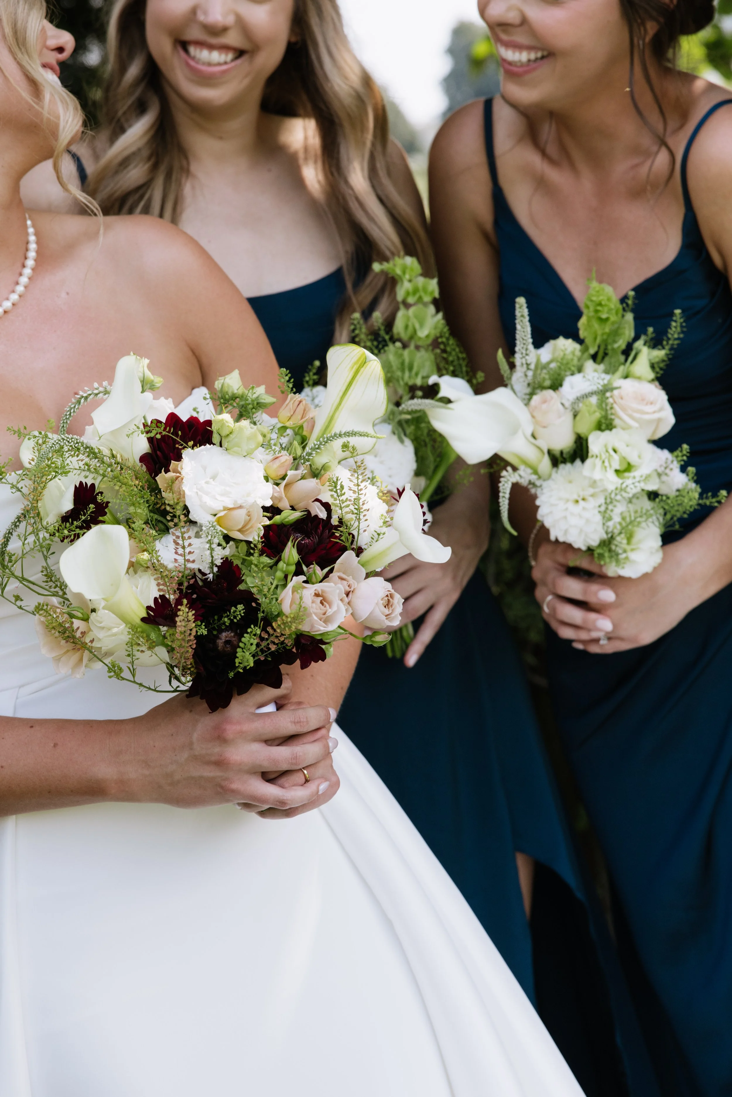 Close-up of three women holding bouquets of white, blush, and dark red flowers, smiling and wearing dark blue dresses, outdoors on a sunny day.