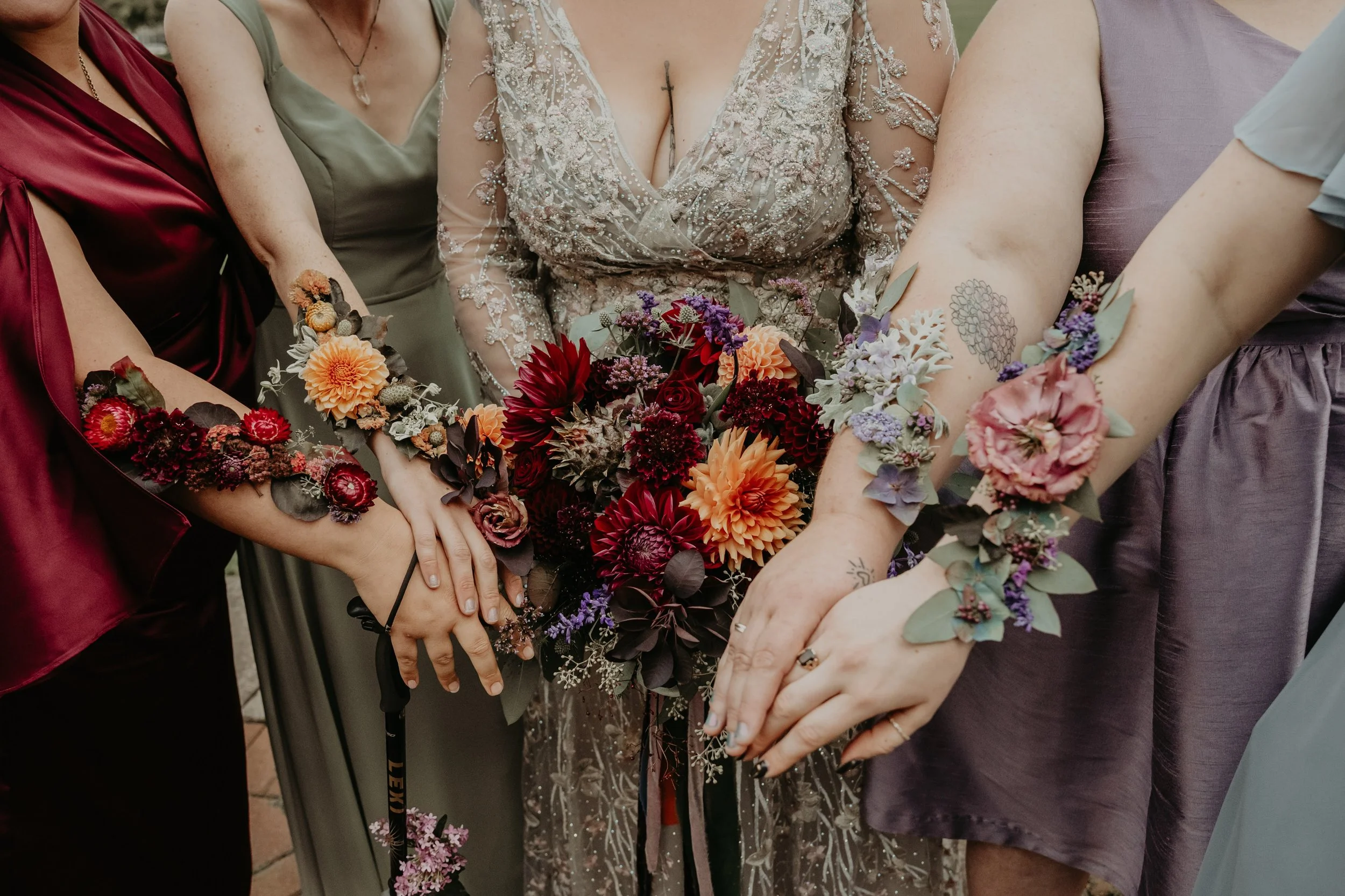 Group of women at a wedding, with hands resting on a large bouquet of orange, red, purple, and white flowers, wearing elegant dresses with floral tattoos.