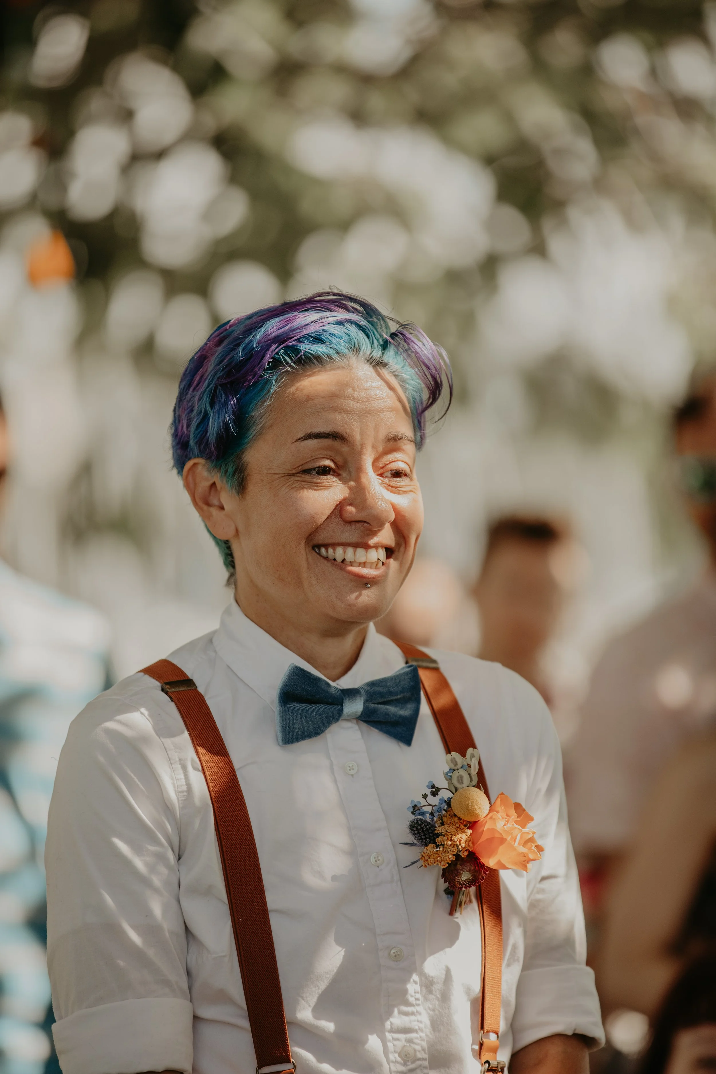 A smiling person with colorful, rainbow hair, wearing a white shirt with rolled-up sleeves, a blue bow tie, orange suspenders, and a floral boutonniere, in an outdoor setting with blurred background.