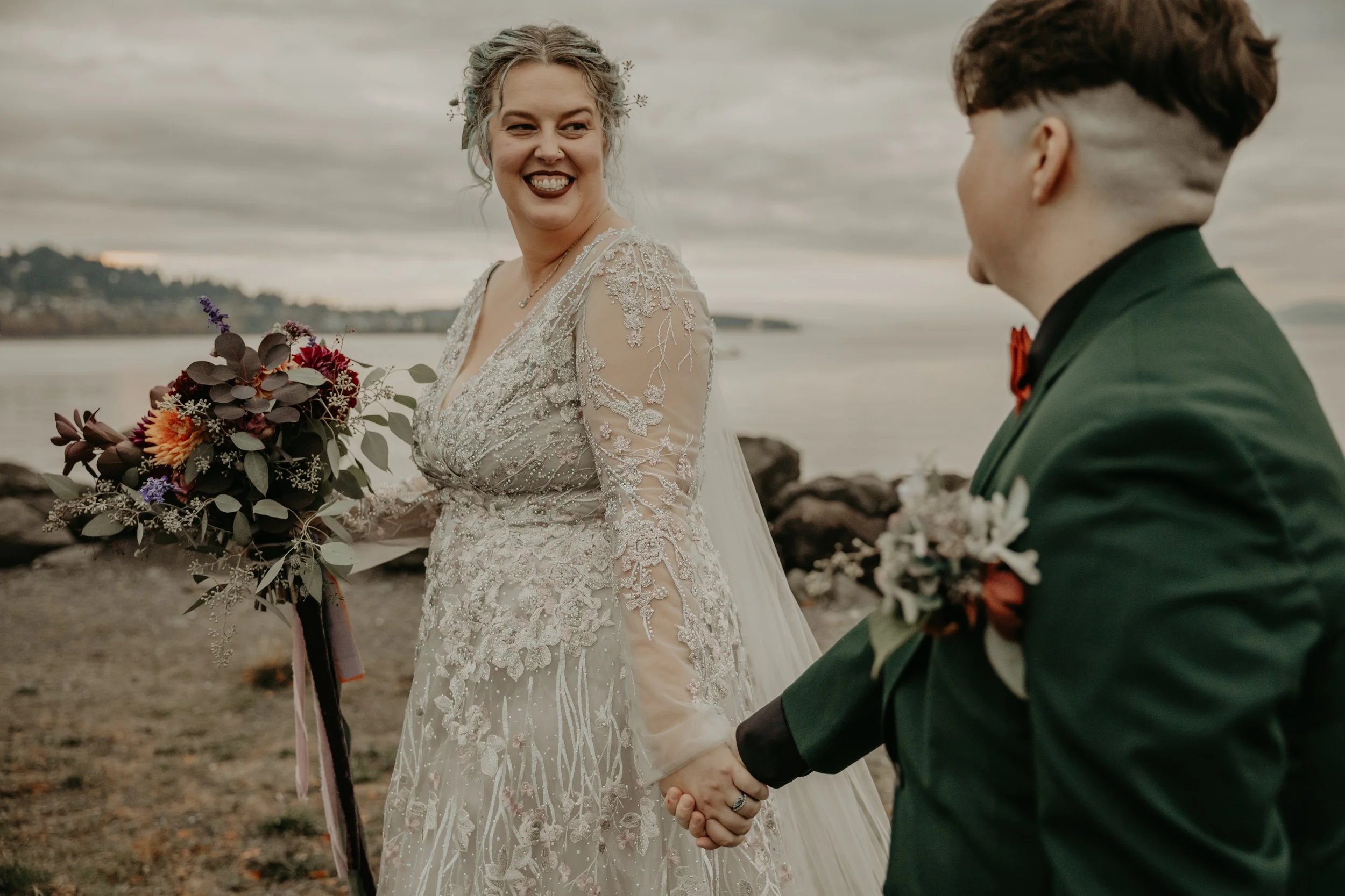 A bride and groom holding hands outdoors, with the bride smiling and looking at the groom, near a body of water and rocky shoreline.