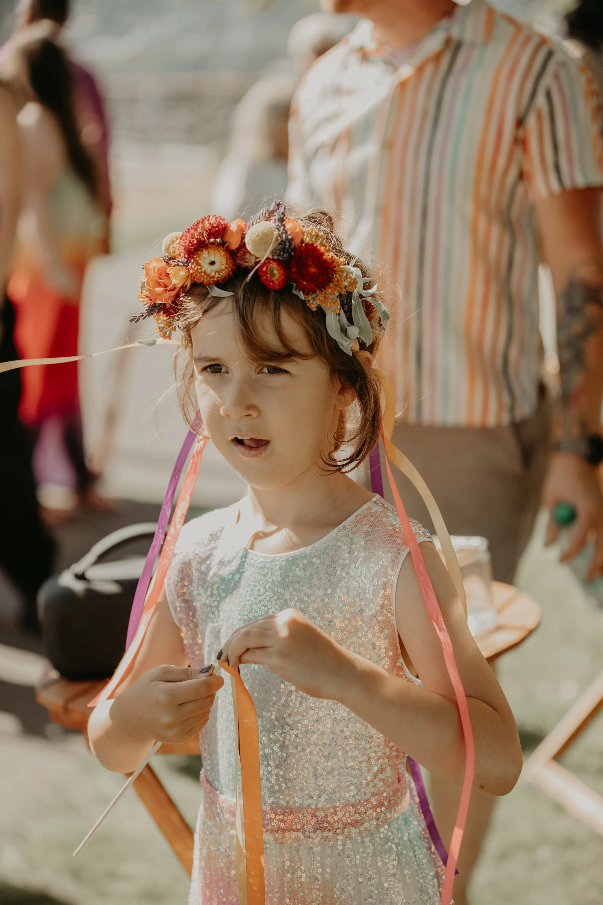 A young girl wearing a sparkly dress and a colorful flower crown for a Pride wedding, outdoors during daylight, with a curious expression on her face.