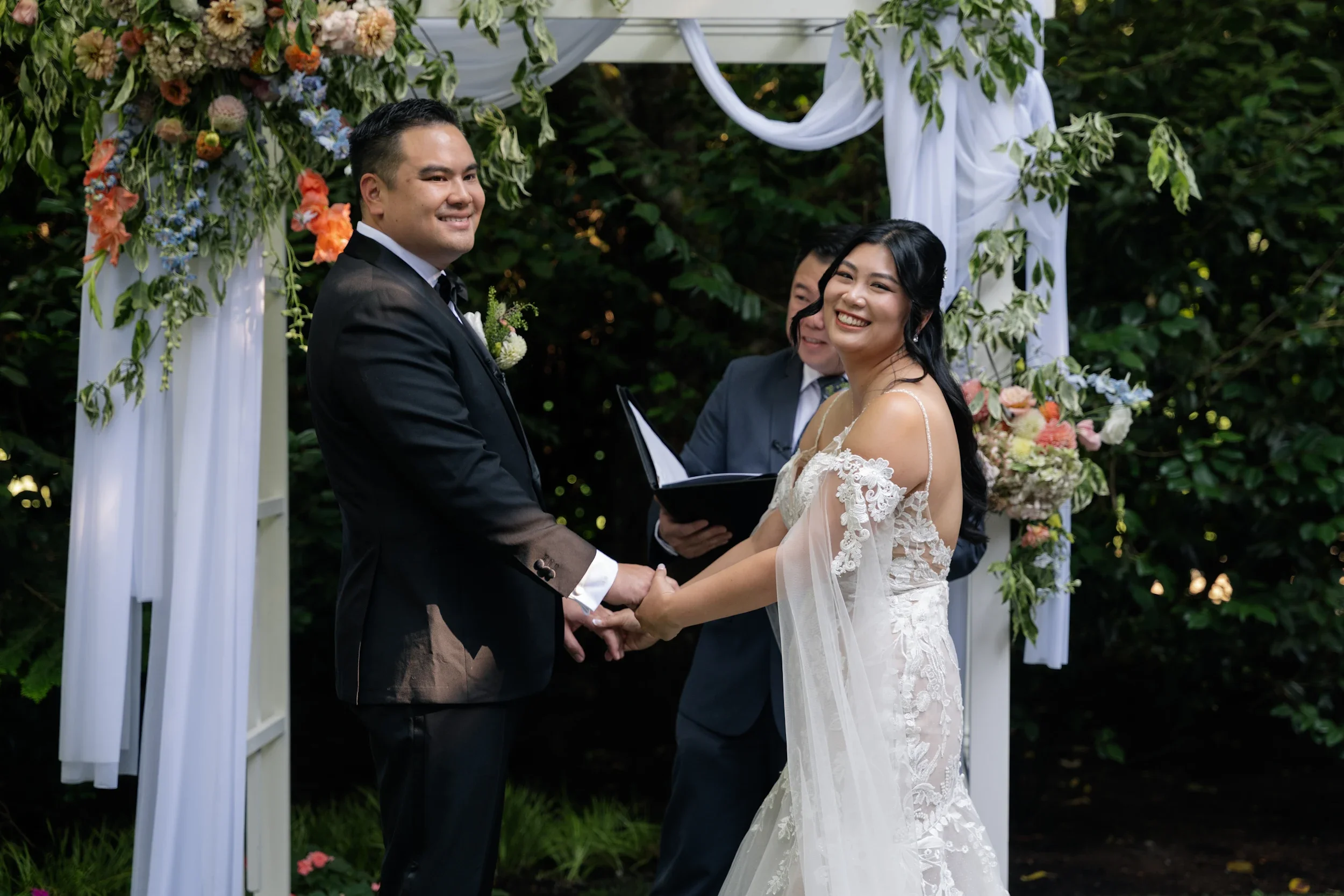 A wedding couple holding hands and smiling at each other during their outdoor wedding ceremony, with an officiant standing behind them under a wedding arch decorated with flowers and white fabric, surrounded by greenery.
