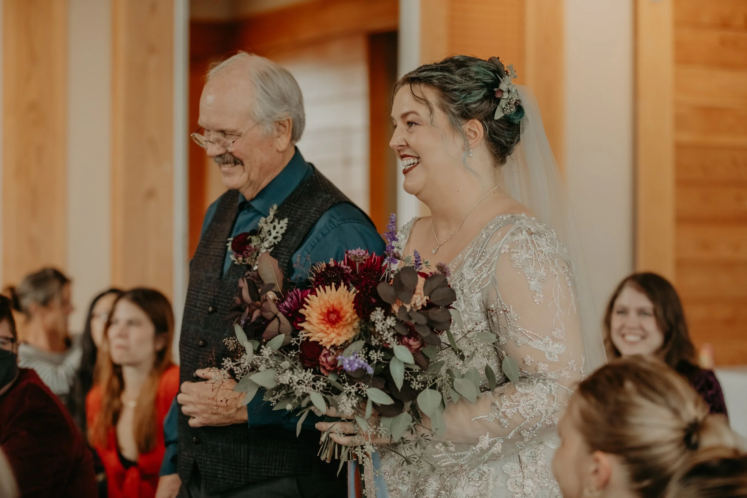 A bride walking down the aisle with her father, holding a bouquet of flowers, smiling happily, inside a wooden-paneled room with seated guests in the background.