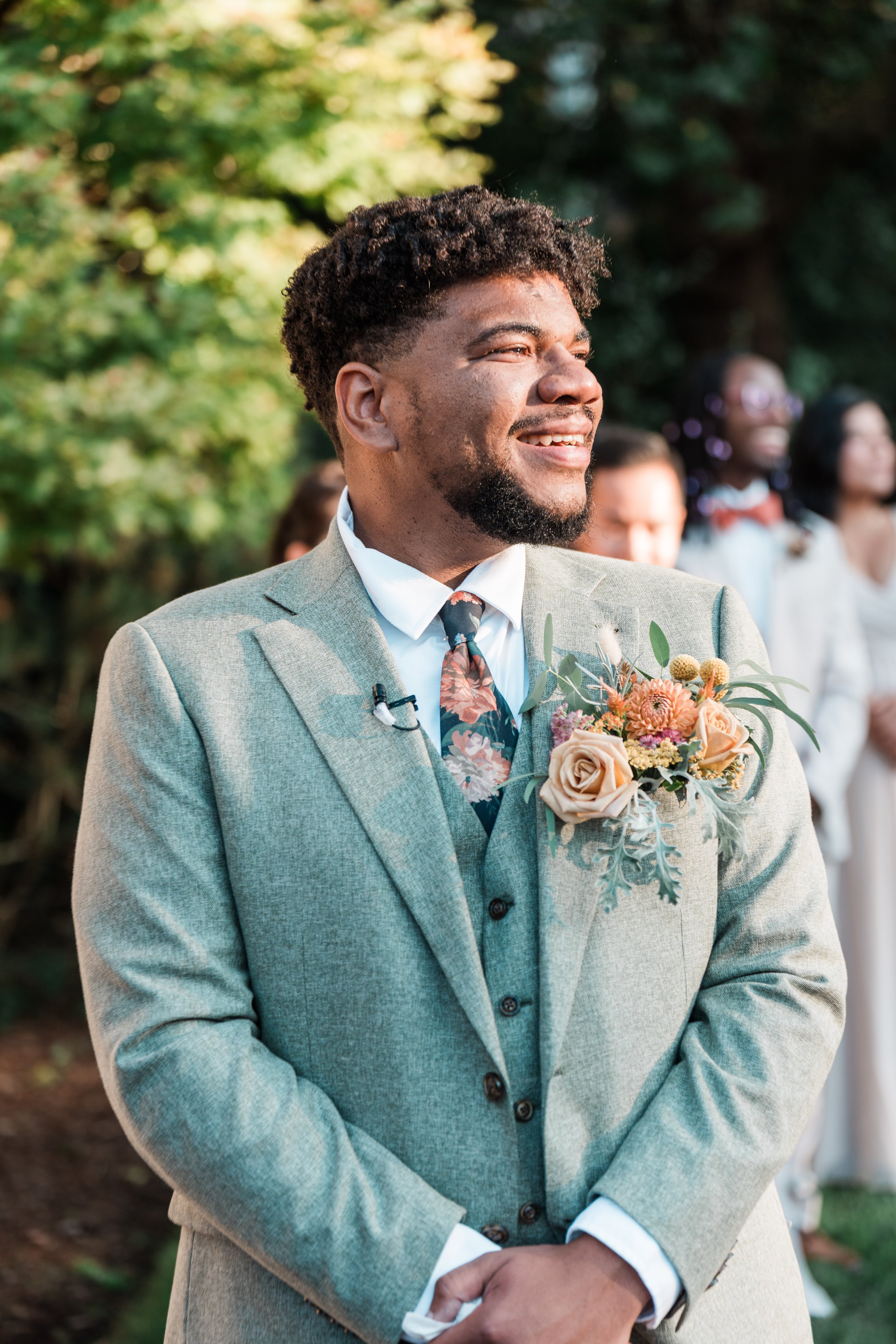 A smiling man in a light gray suit with a floral tie and a boutonniere of flowers on his lapel, standing outdoors during a wedding ceremony.