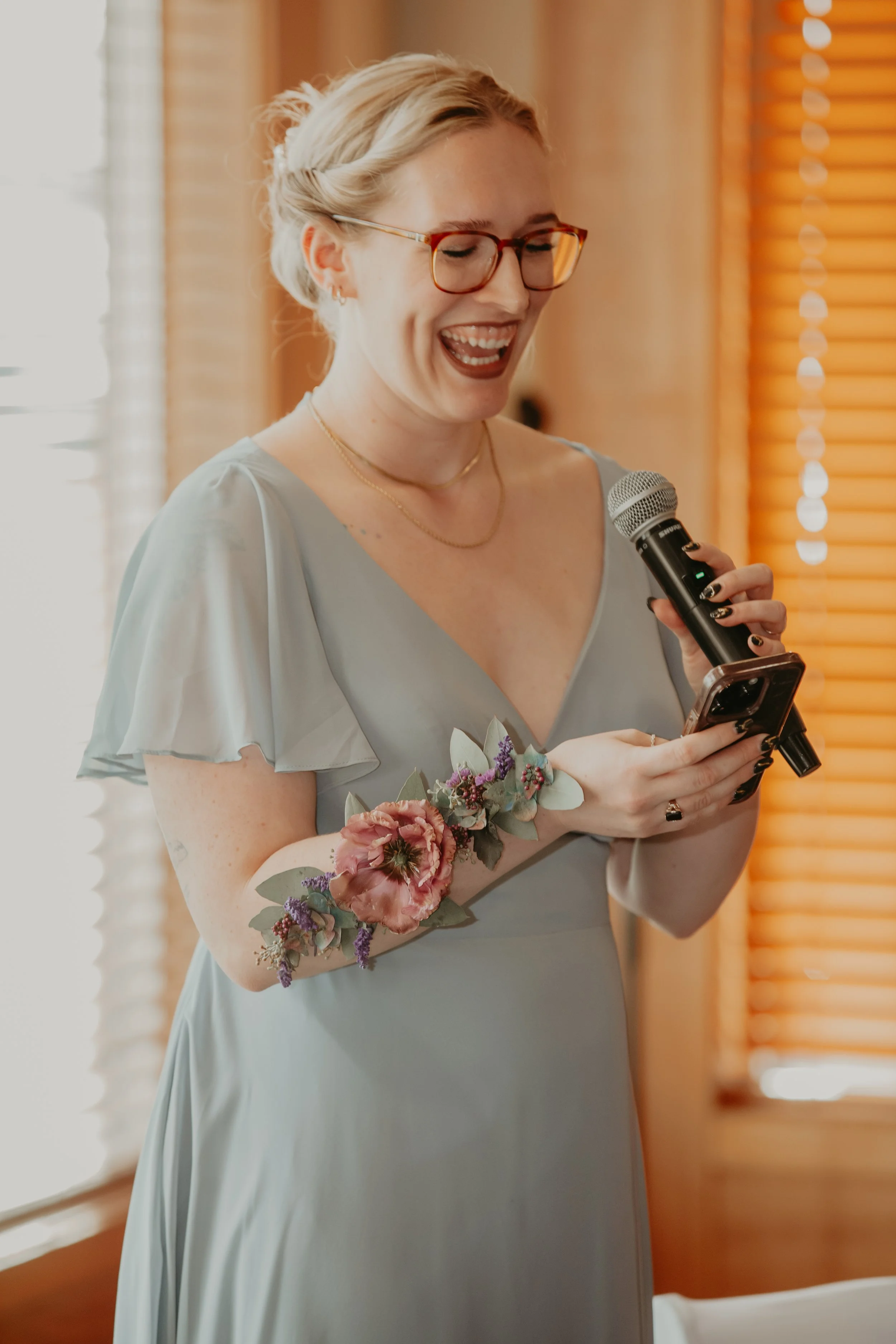 A bridesmaid with blonde hair, glasses, and a floral tattoo is smiling, holding a microphone in one hand and a phone in the other, indoors with wooden blinds in the background.
