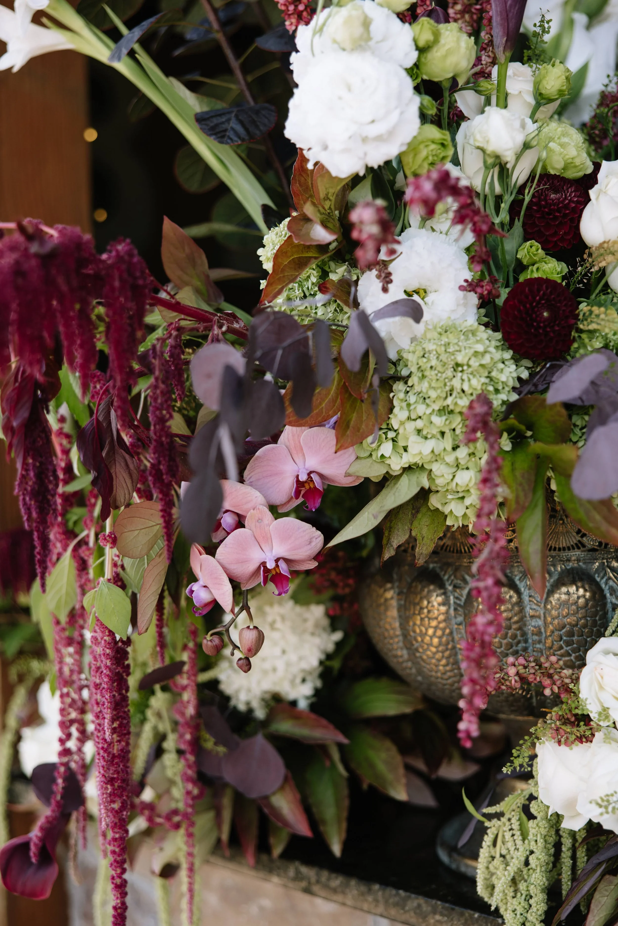 A colorful wedding arrangement of various flowers including white, pink, and burgundy blooms, with green and dark purple foliage, in a decorative metallic vase.