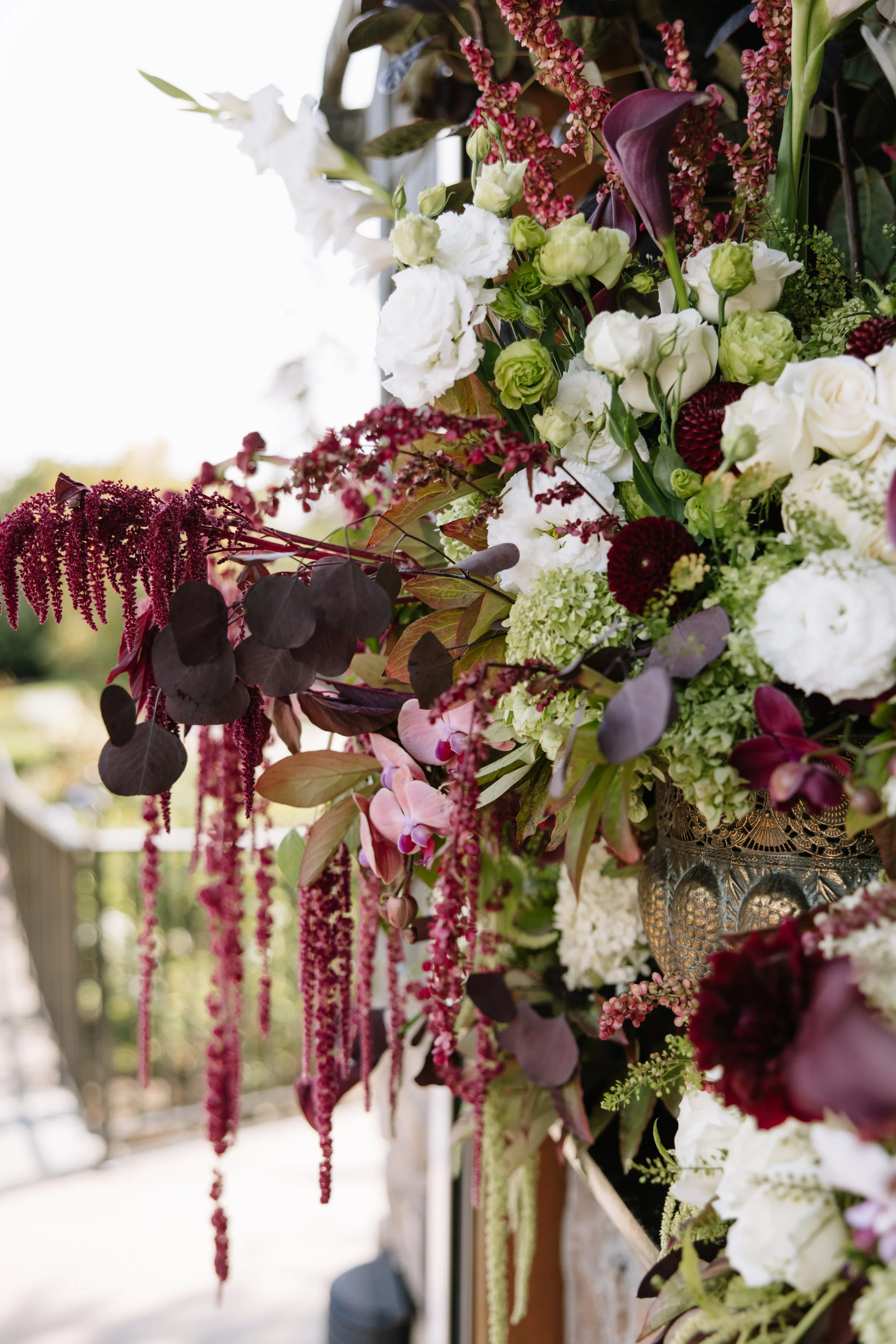 Close-up of a lush wedding floral arrangement with white, purple, and green flowers, including roses, Calla lilies, and orchids, accented with dark purple and maroon foliage, hanging on a railing outdoors.