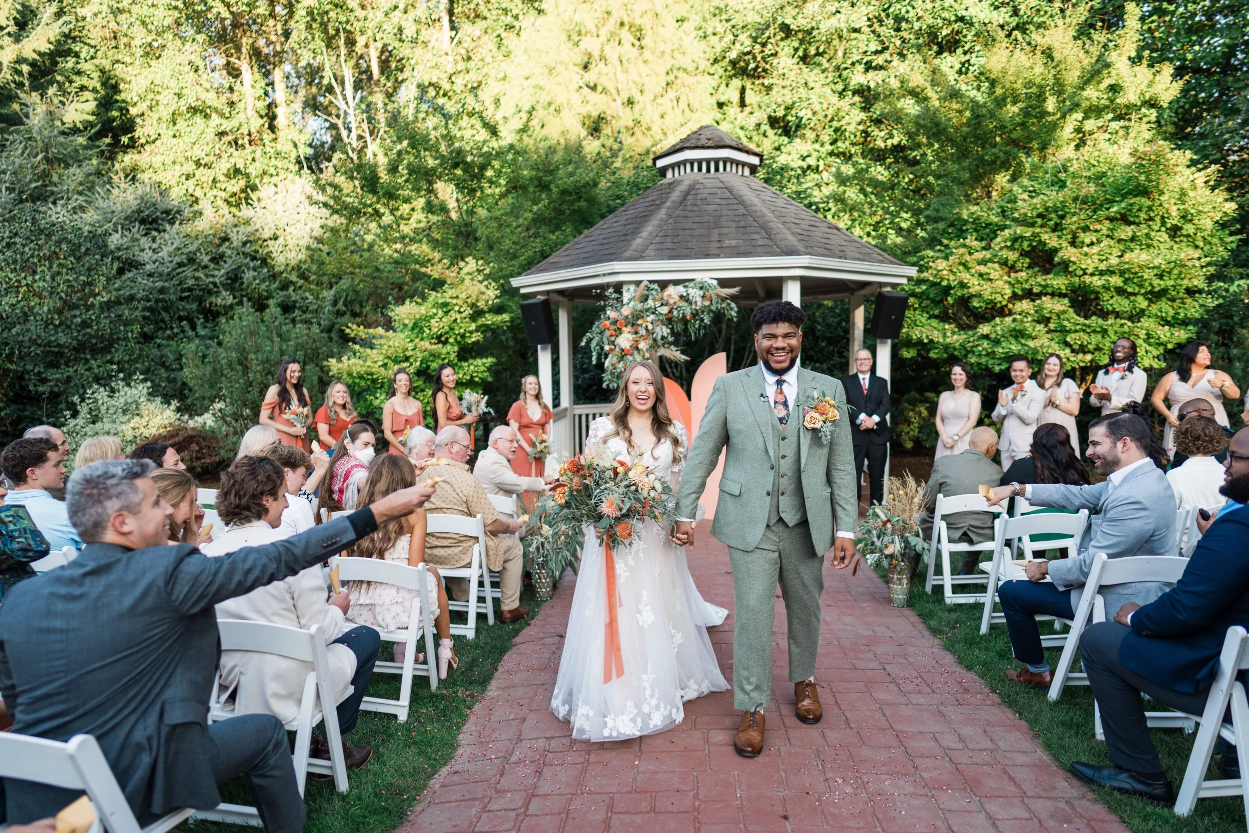 A couple, the bride and groom, smiling and holding hands while walking down the aisle at their outdoor wedding ceremony, surrounded by guests seated on white chairs and a lush green background.