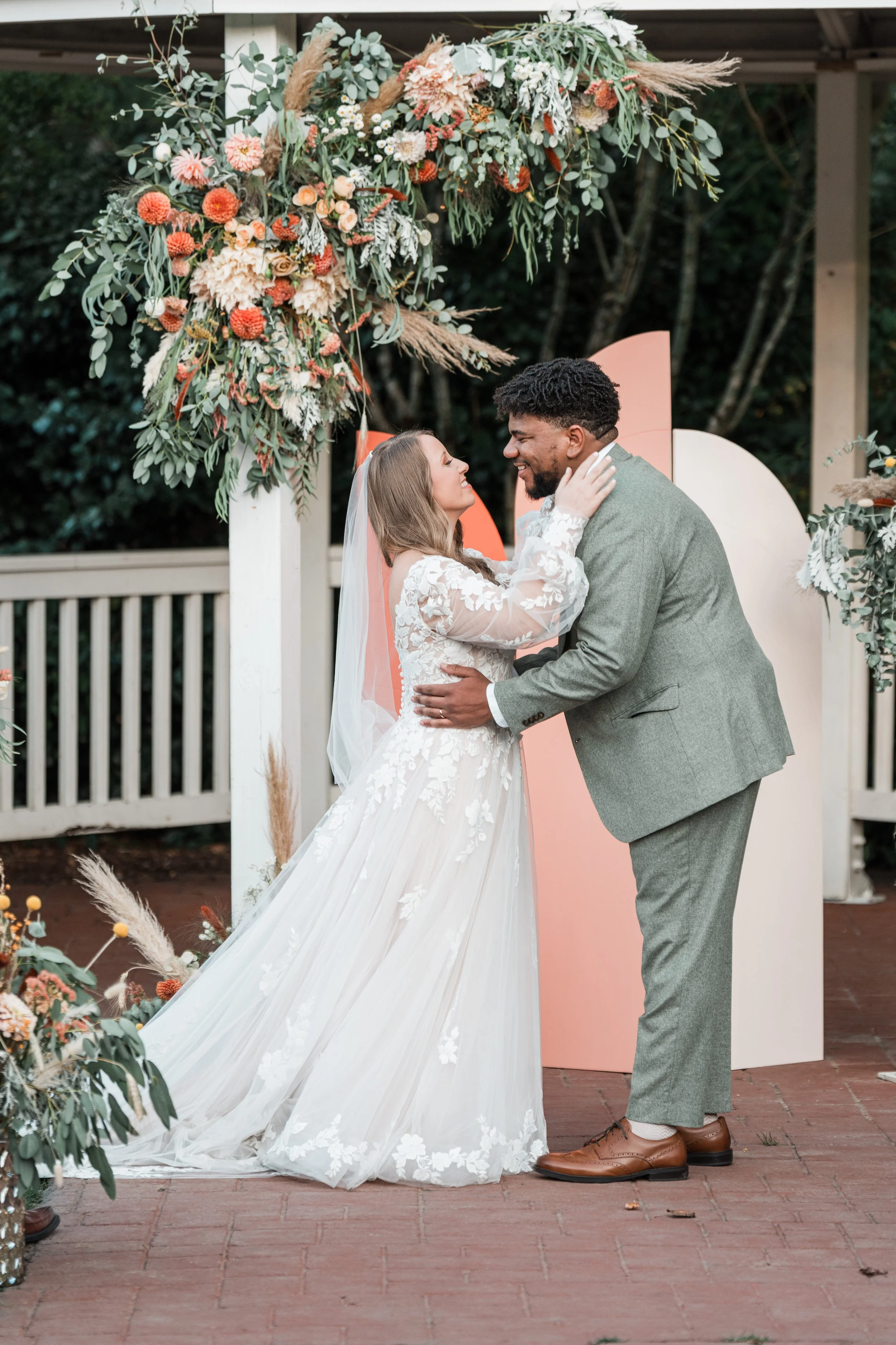 A bride and groom share a loving moment during their wedding ceremony outdoors, under a floral arch decorated with pink, orange, and white flowers and greenery.