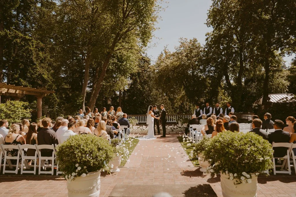 Outdoor wedding ceremony with bride and groom exchanging vows, seated guests on both sides, officiant, and wedding party in the background, surrounded by trees and sunlight.