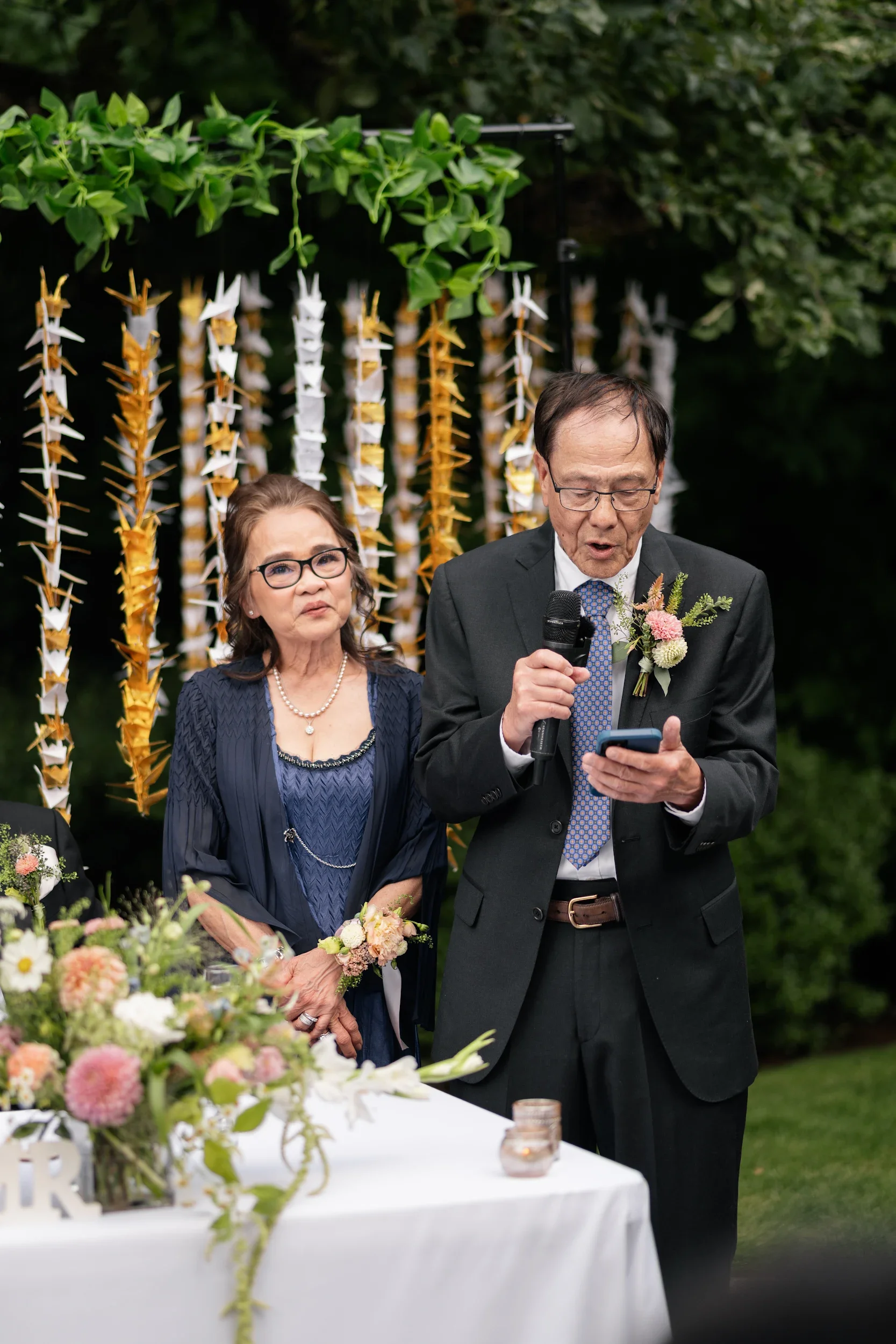An elderly couple at a wedding or formal event outdoors, with the man reading from a phone and holding a microphone, and the woman standing beside him with flowers on her wrist, in front of hanging decorations made of paper or fabric in a garden sett