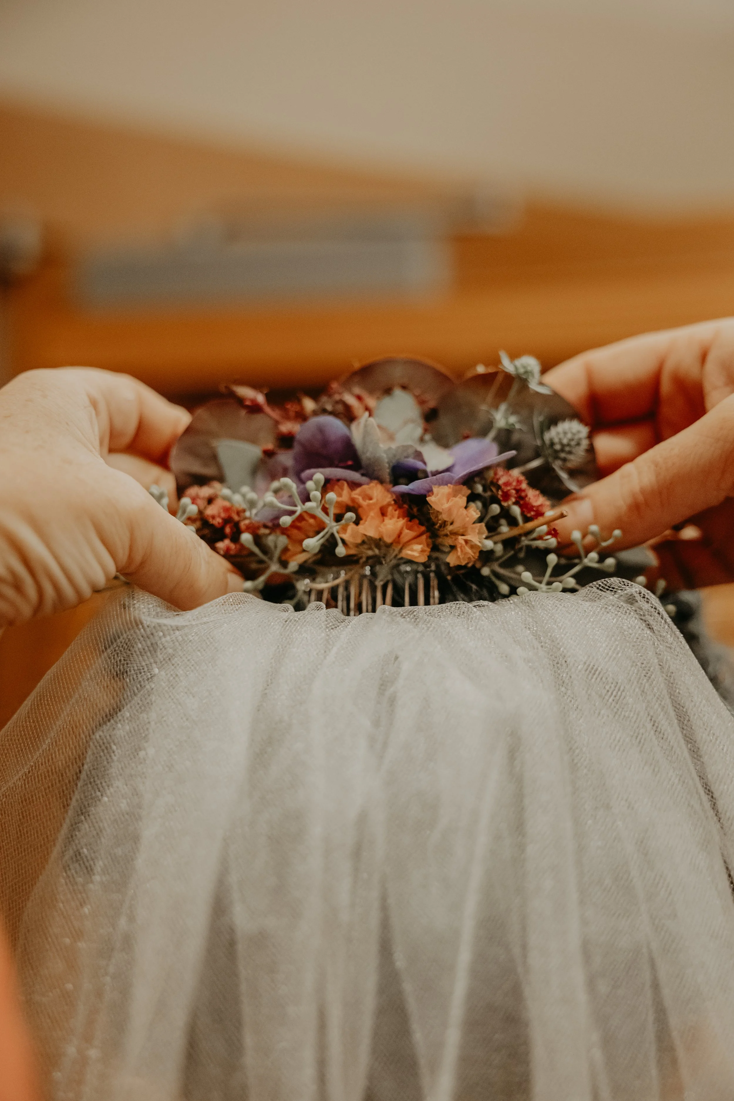 Hands holding a decorative hair comb with flowers and greenery, with a tulle fabric in the foreground.