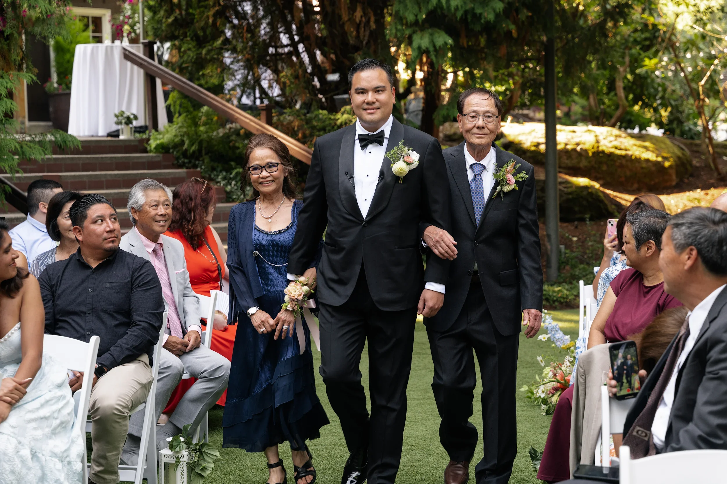 A groom walking arm-in-arm with his parents at an outdoor wedding ceremony holding bouquets, surrounded by seated guests.