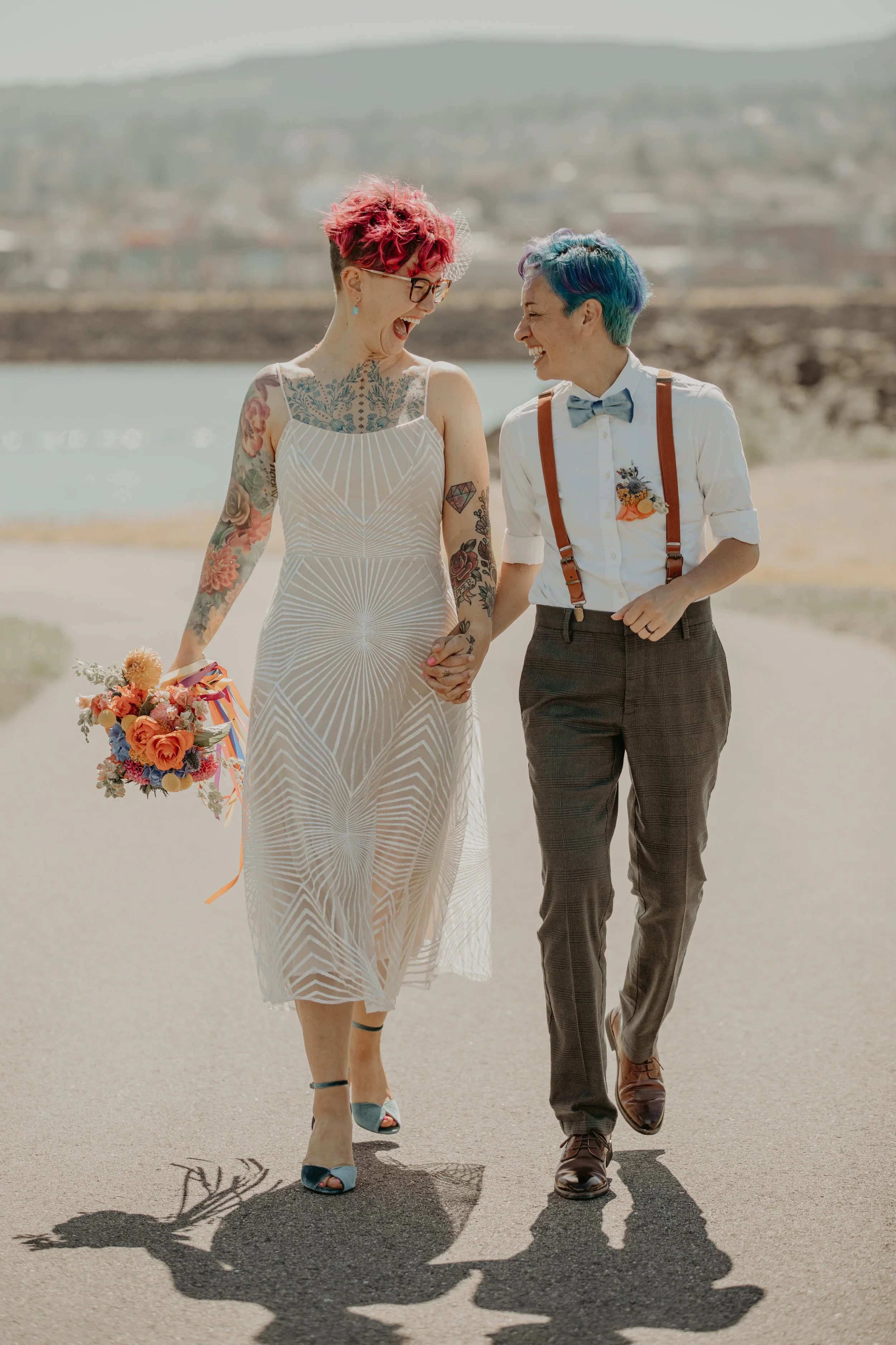 Two women with colorful hair celebrating outdoors, holding hands, with a lake and mountains in the background.