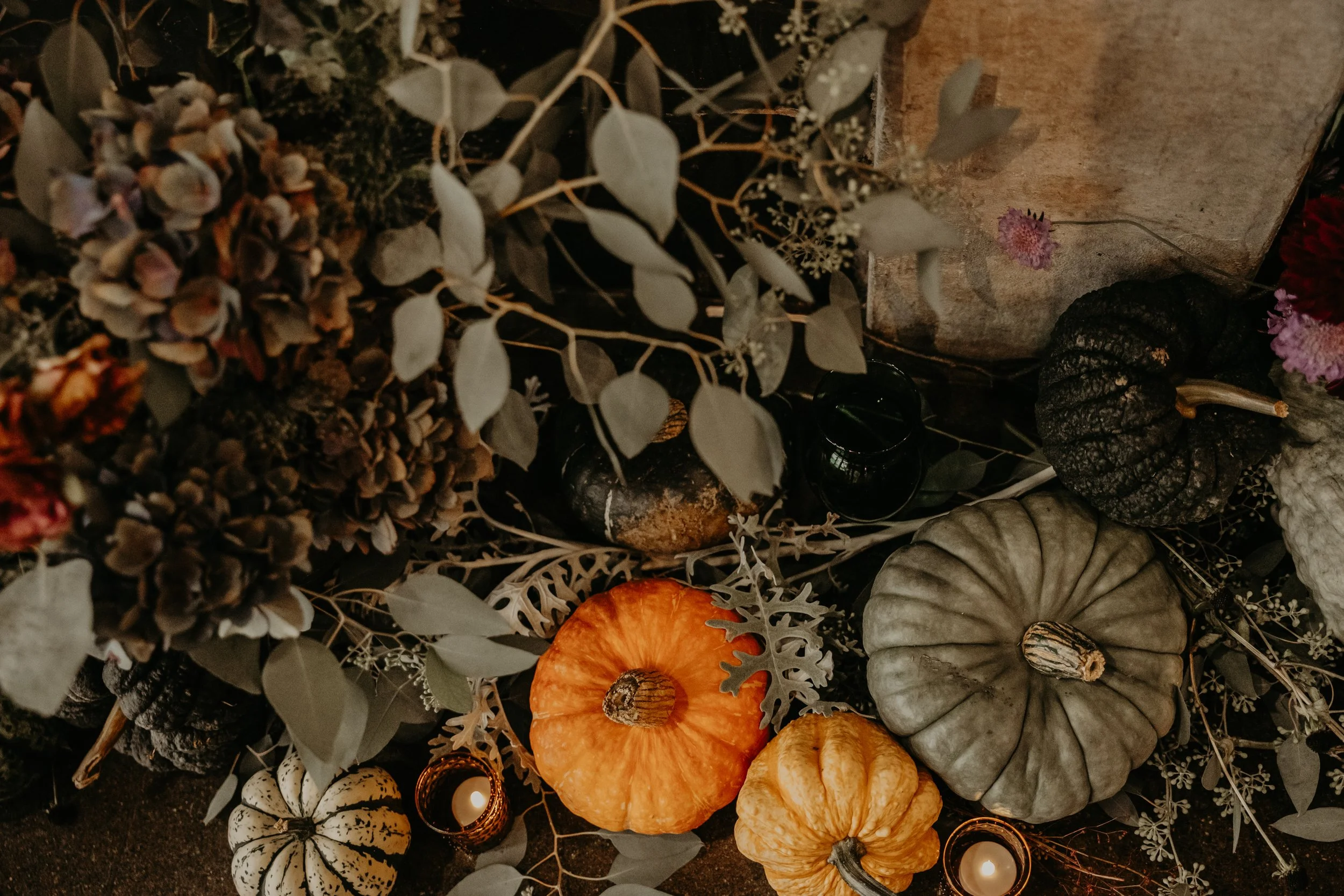 Wedding installation for autumn of pumpkins, gourds, dried flowers, candles, and foliage arranged on dark surface