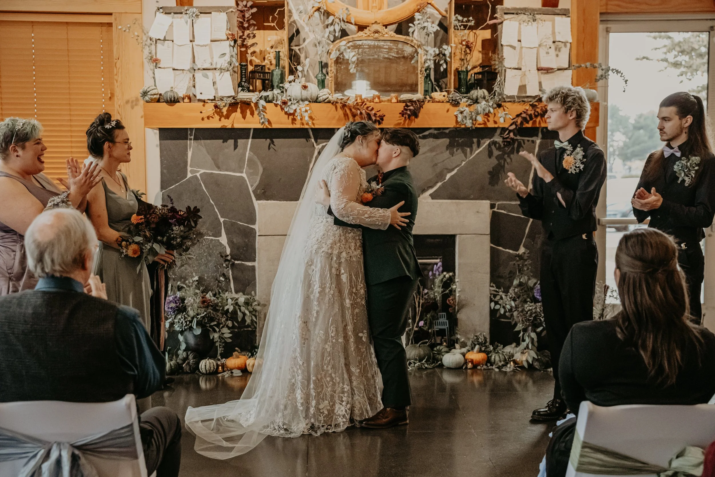 A couple kissing during their wedding ceremony in front of a fireplace, surrounded by friends and family, with autumn-themed decorations.