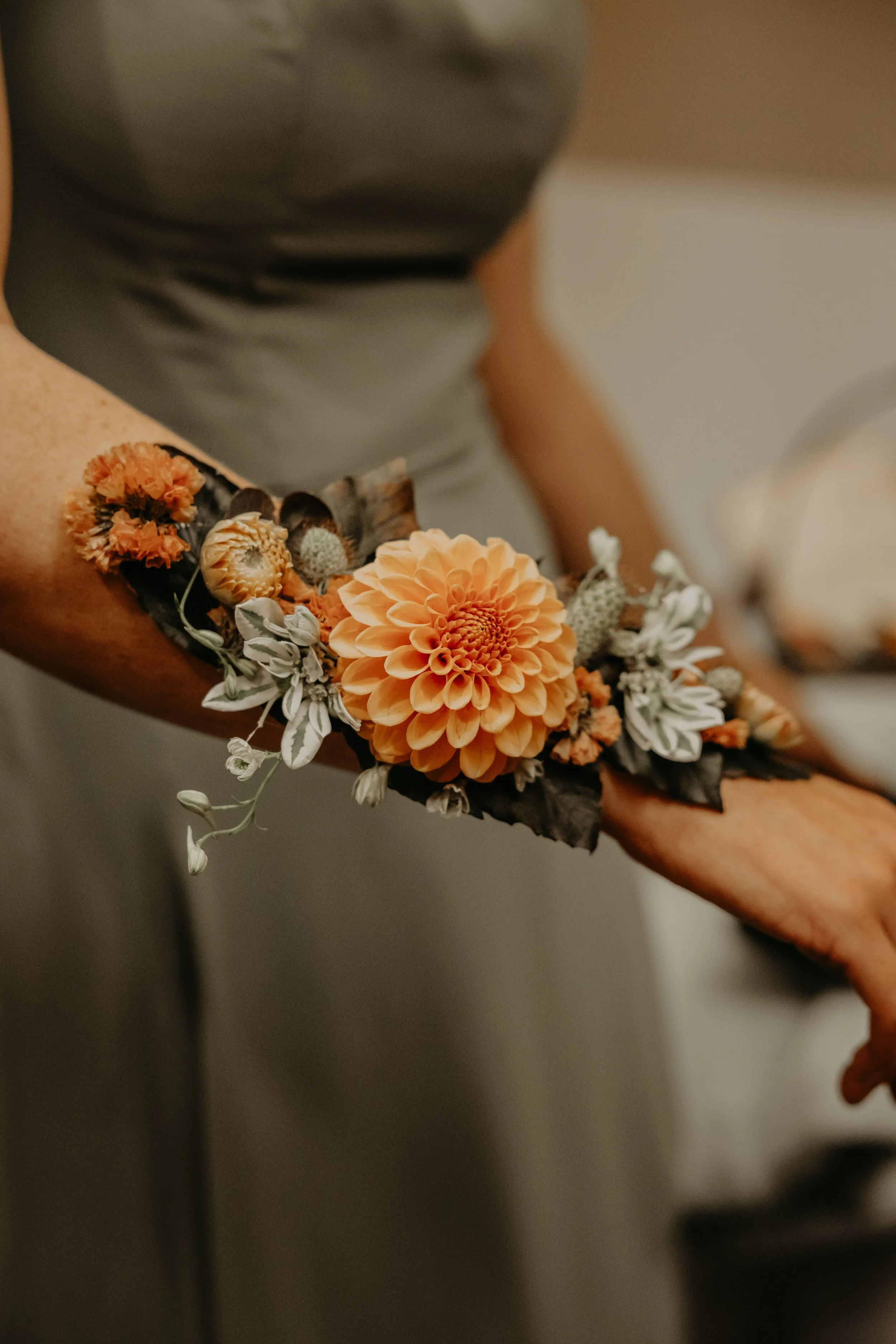 A person's arm with a floral tattoo, featuring an orange dahlia and smaller flowers and greenery, is shown.