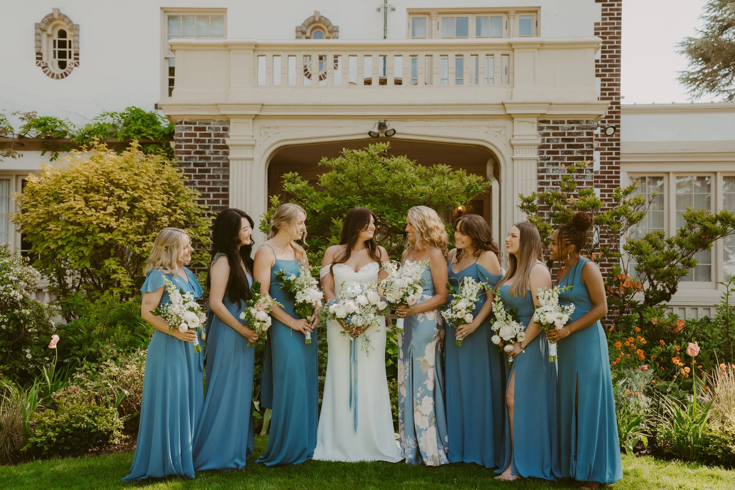 A bride and her bridesmaids standing outdoors in front of a house with garden plants, all holding bouquets, dressed in blue and floral dresses.
