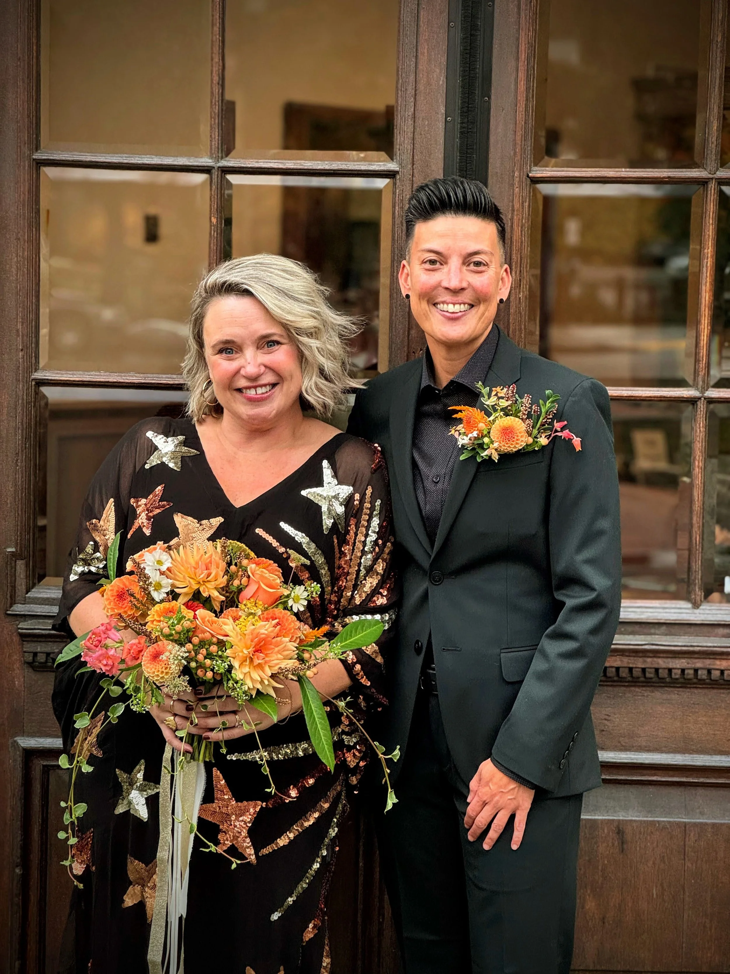 Two smiling women standing in front of a wooden door with glass panes, one holding a bouquet of orange and pink flowers, both dressed in formal attire with floral accents.