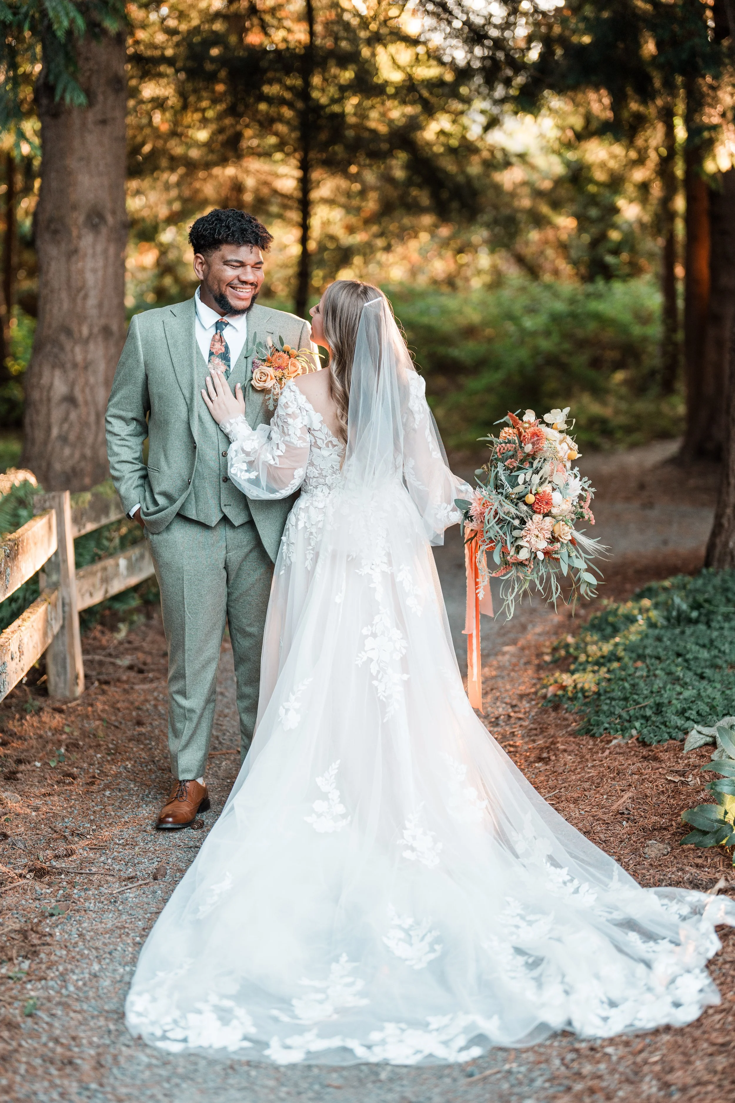 A bride and groom smiling and looking at each other outdoors in a wooded area during their wedding, with the bride holding a colorful bouquet and wearing a long white dress with lace details, and the groom wearing a light gray suit with a floral tie.
