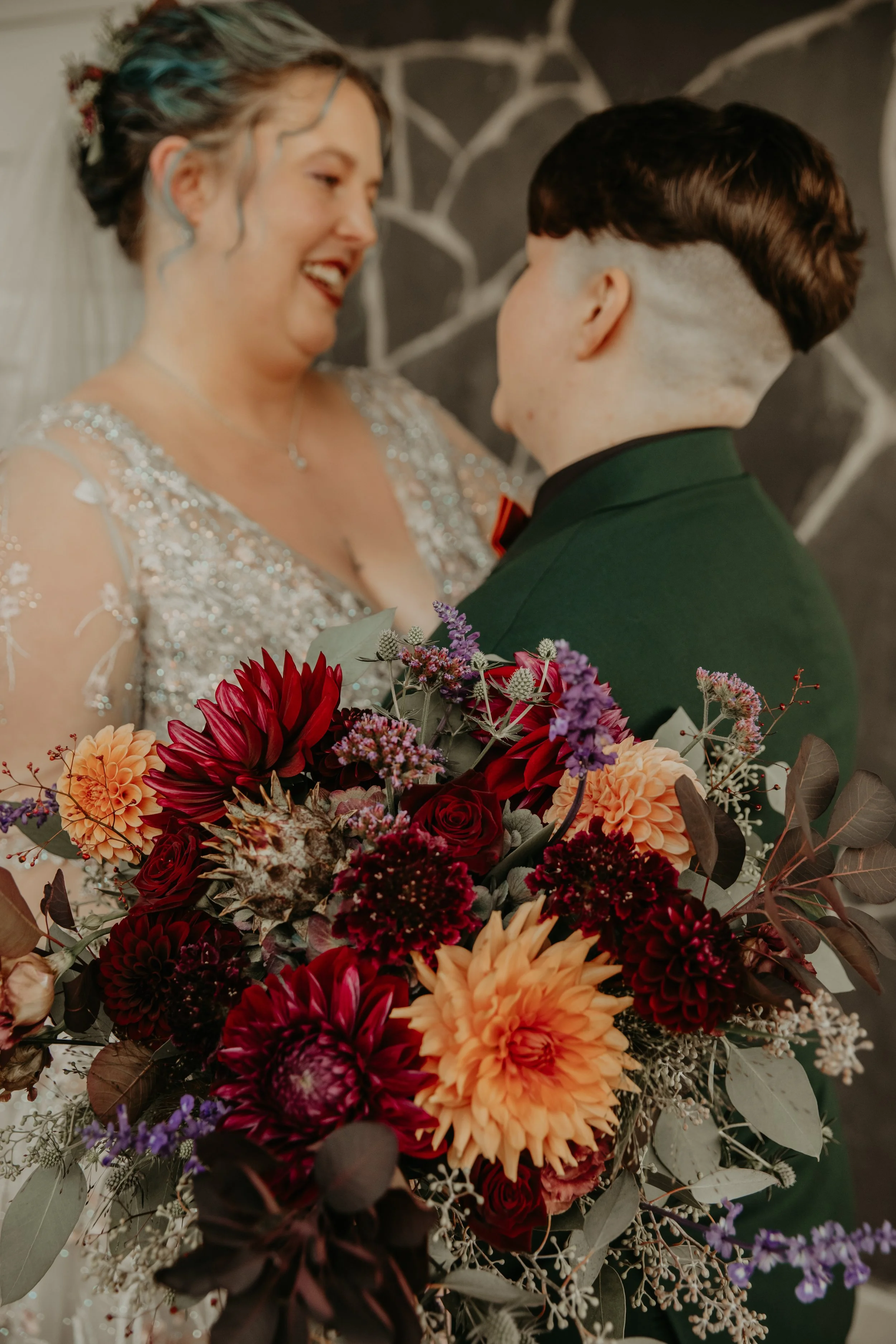 A bride and groom sharing a moment, with the bride holding a large bouquet of red, orange, purple, and dark red flowers, in front of a dark, patterned wall.