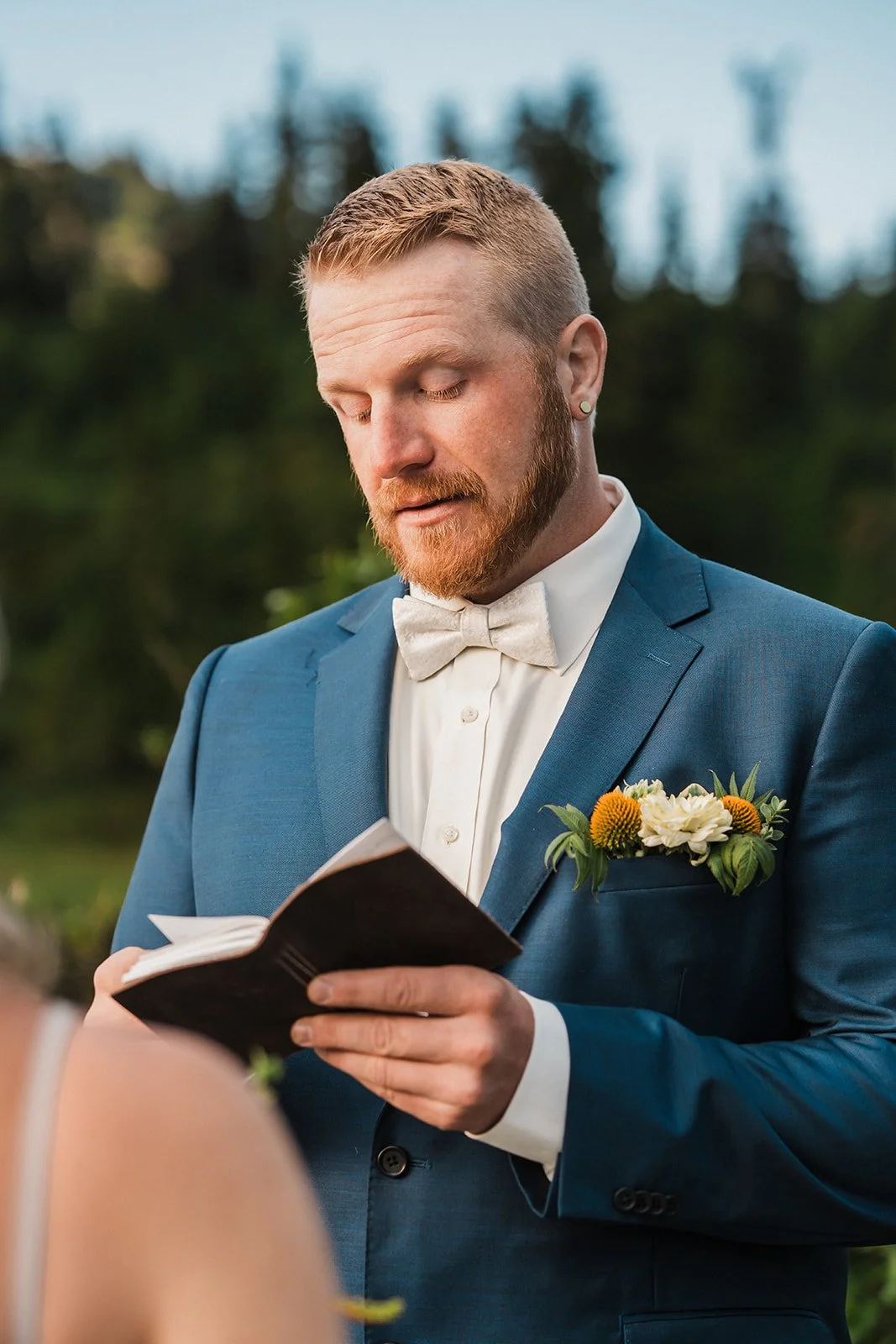 A groom in a blue suit with a white bow tie and boutonniere reading vows during an outdoor wedding ceremony.