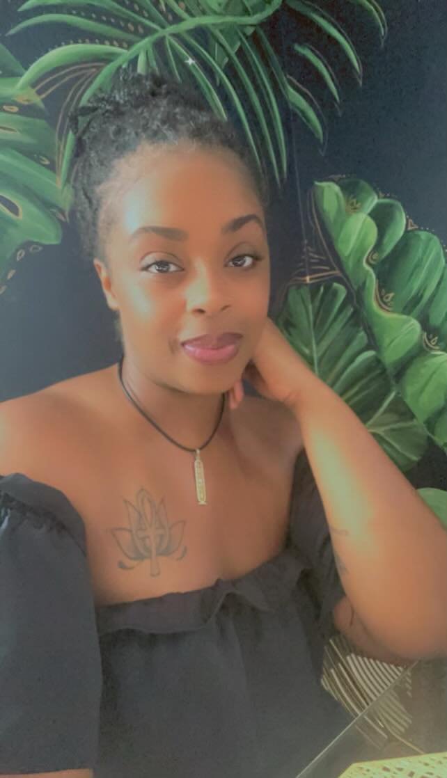 A woman with braided hair and makeup, wearing a black off-the-shoulder top, a necklace with a gold pendant, and a chest tattoo of a lotus flower with a line in the middle, posing in front of green tropical leaves.