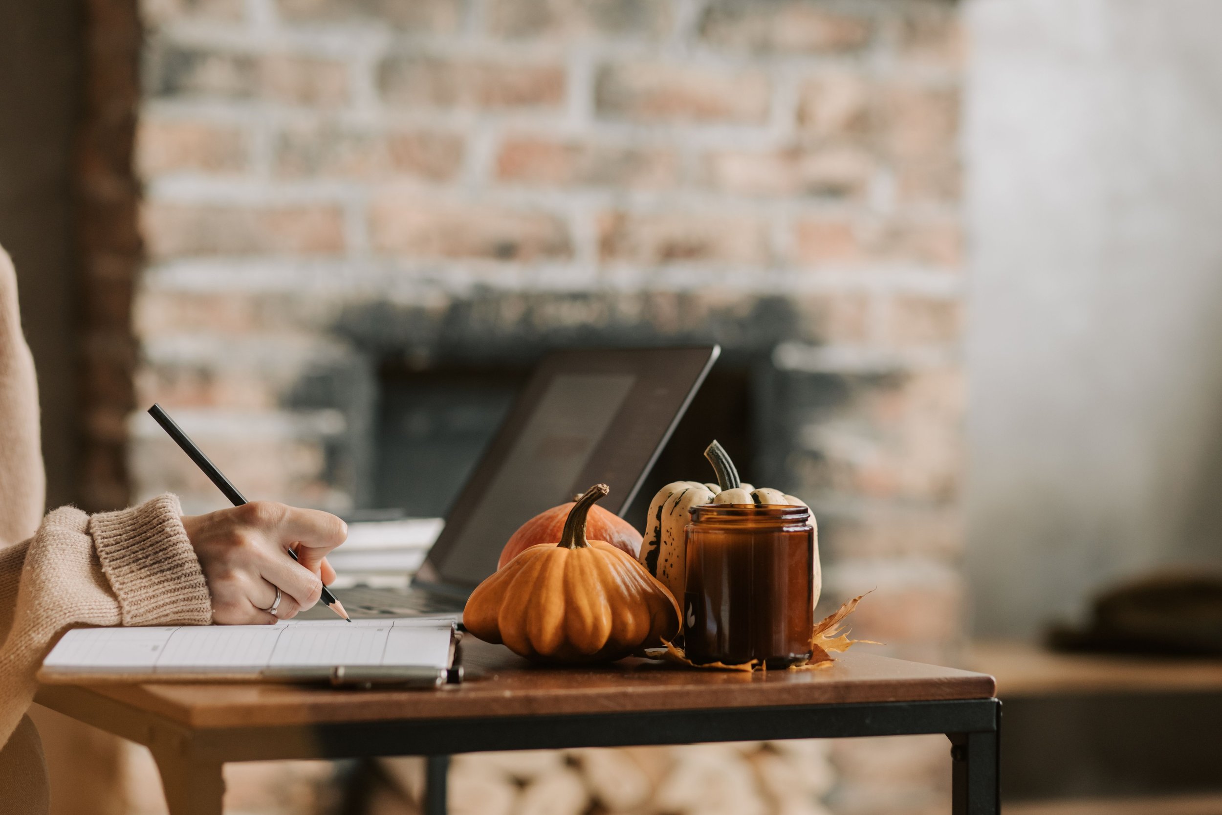 Person writing in a notebook beside a laptop, small pumpkins and a candle in front of a fireplace.