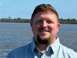 A man with short brown hair and a beard, wearing a light blue button-up shirt, standing outdoors near a body of water with trees and a blue sky in the background.