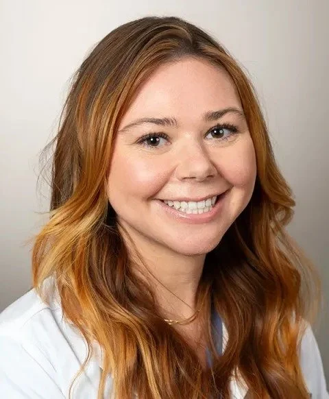 A young woman with long, wavy auburn hair, smiling, wearing a white top, against a plain neutral background.