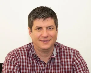 A man with short dark hair, wearing a red and white checkered shirt, smiling at the camera against a plain light background.