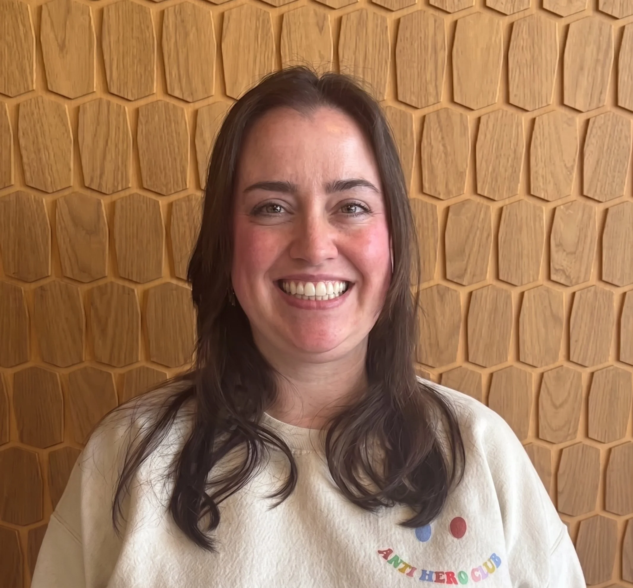 Close-up of a woman smiling, with long dark hair, standing in front of a honeycomb-style wooden wall, wearing a white shirt with colorful text.