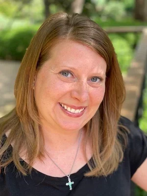 A woman with shoulder-length light brown hair smiling outdoors with greenery in the background.