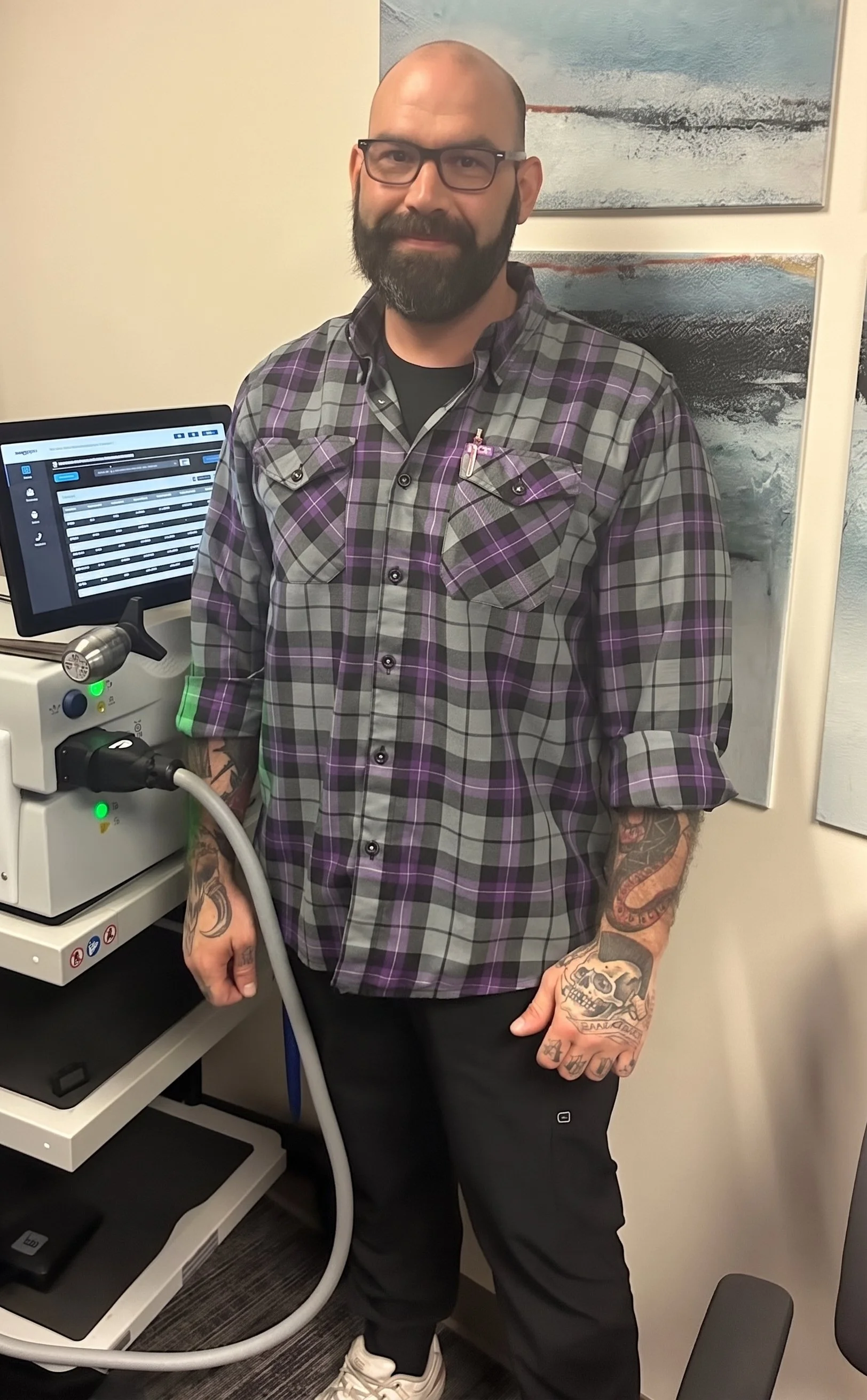 A man with tattoos on his arms, a beard, glasses, and a bald head, is standing next to medical equipment in a room with abstract art on the wall behind him.