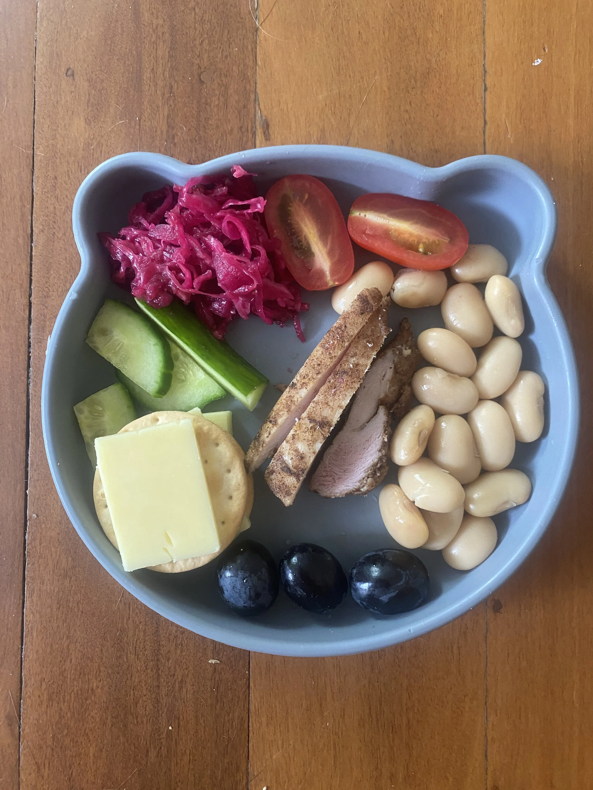 An example of a quick toddler meal using leftovers. The plate shows legumes, sliced cherry tomatoes, pickled red cabbage, cucumber slices, a slice of cheese, a rice cracker, cooked chicken, and three black olives, all on a wooden surface.