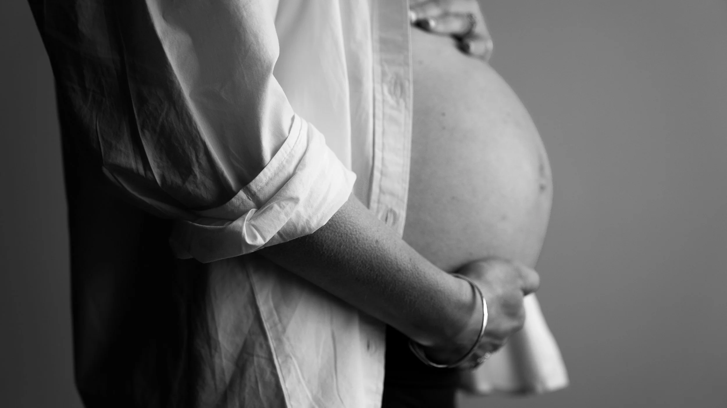Black and white photograph of a pregnant woman wearing a white shirt with rolled-up sleeves, gently cradling her belly with her left hand.