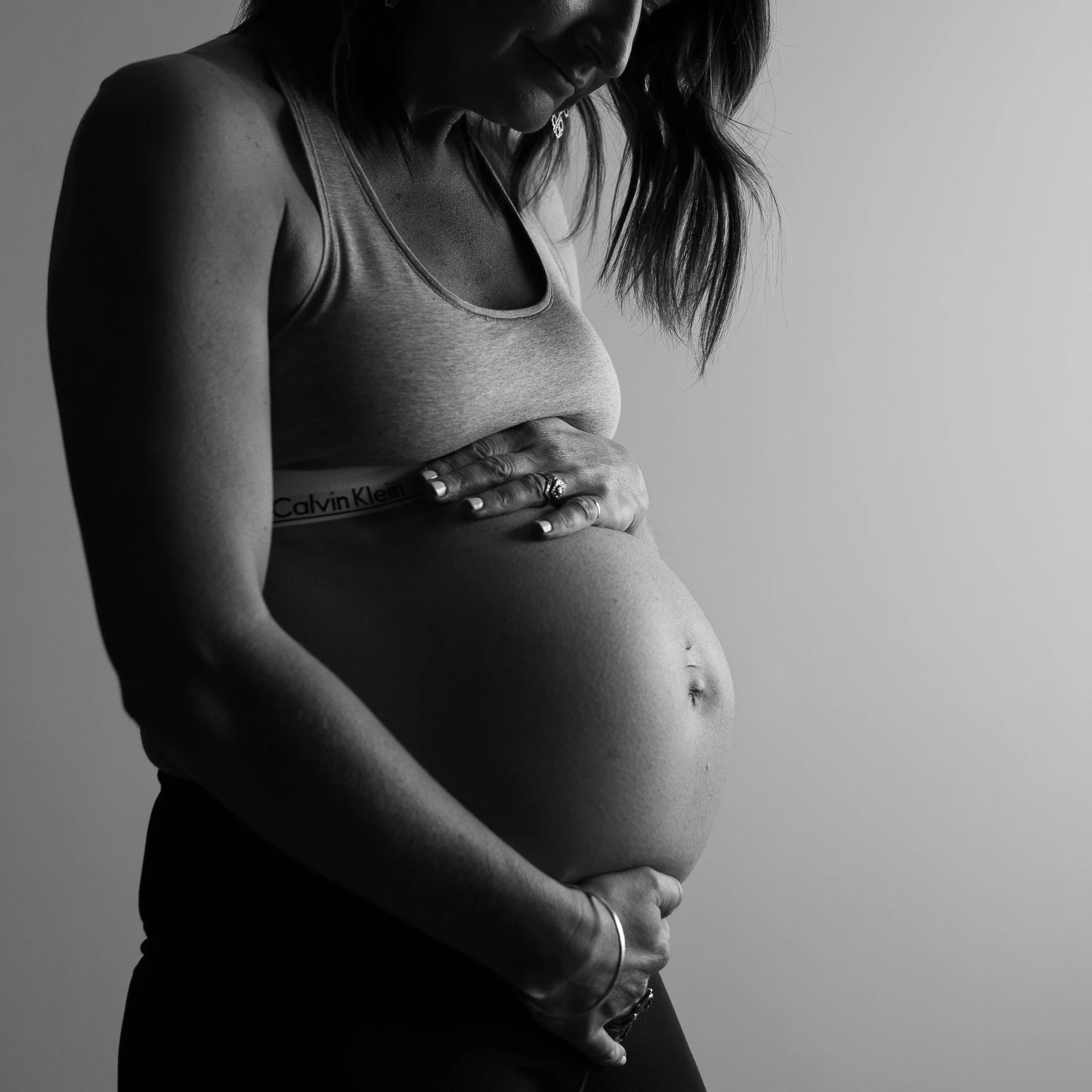 Black and white photo of a pregnant woman in a tank top, gently cradling her belly with both hands, looking down at her stomach.