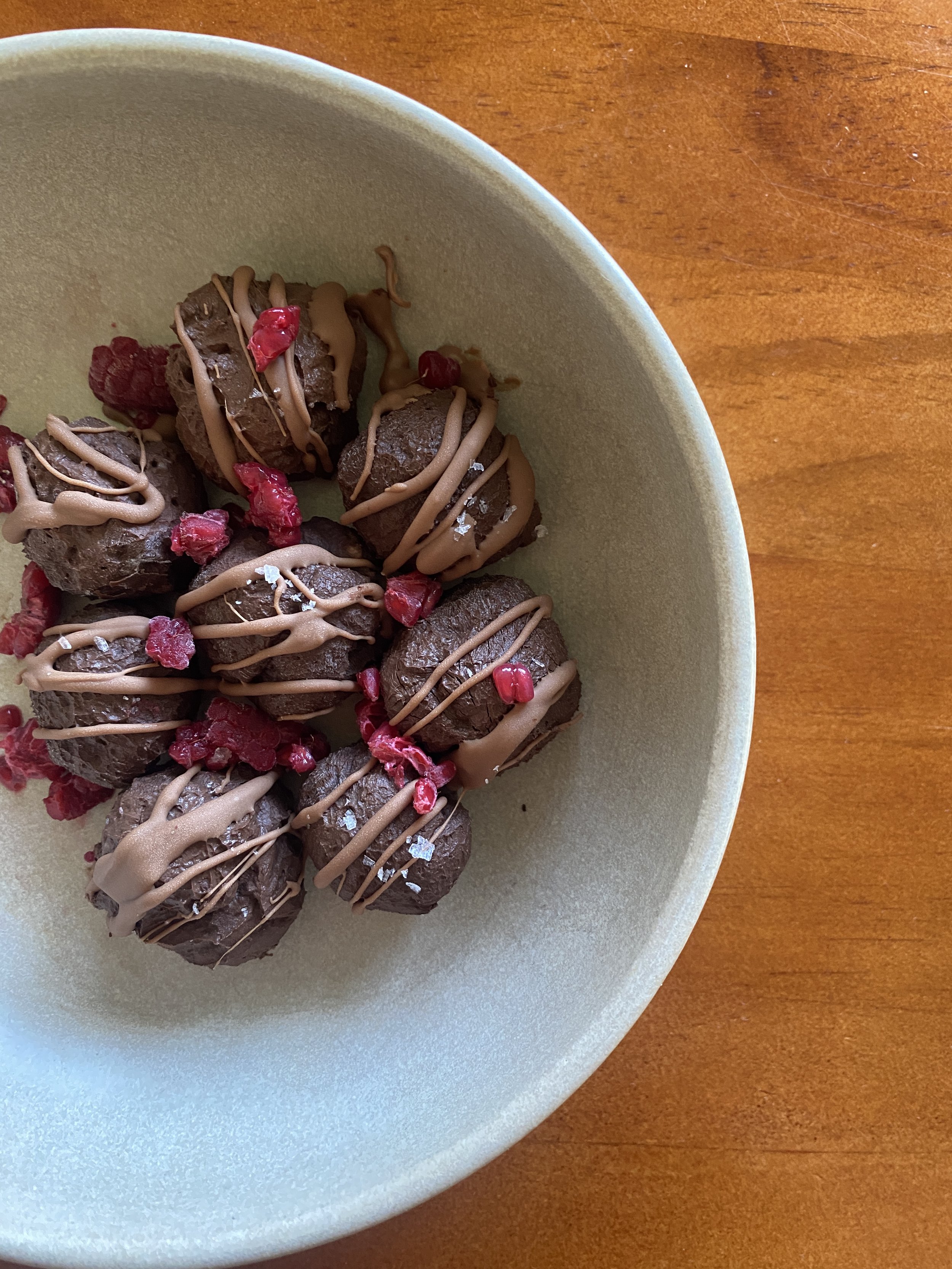 Healthy toddler snack of chocolate protein truffles drizzled with chocolate and topped with red raspberries in a beige bowl on a wooden surface.