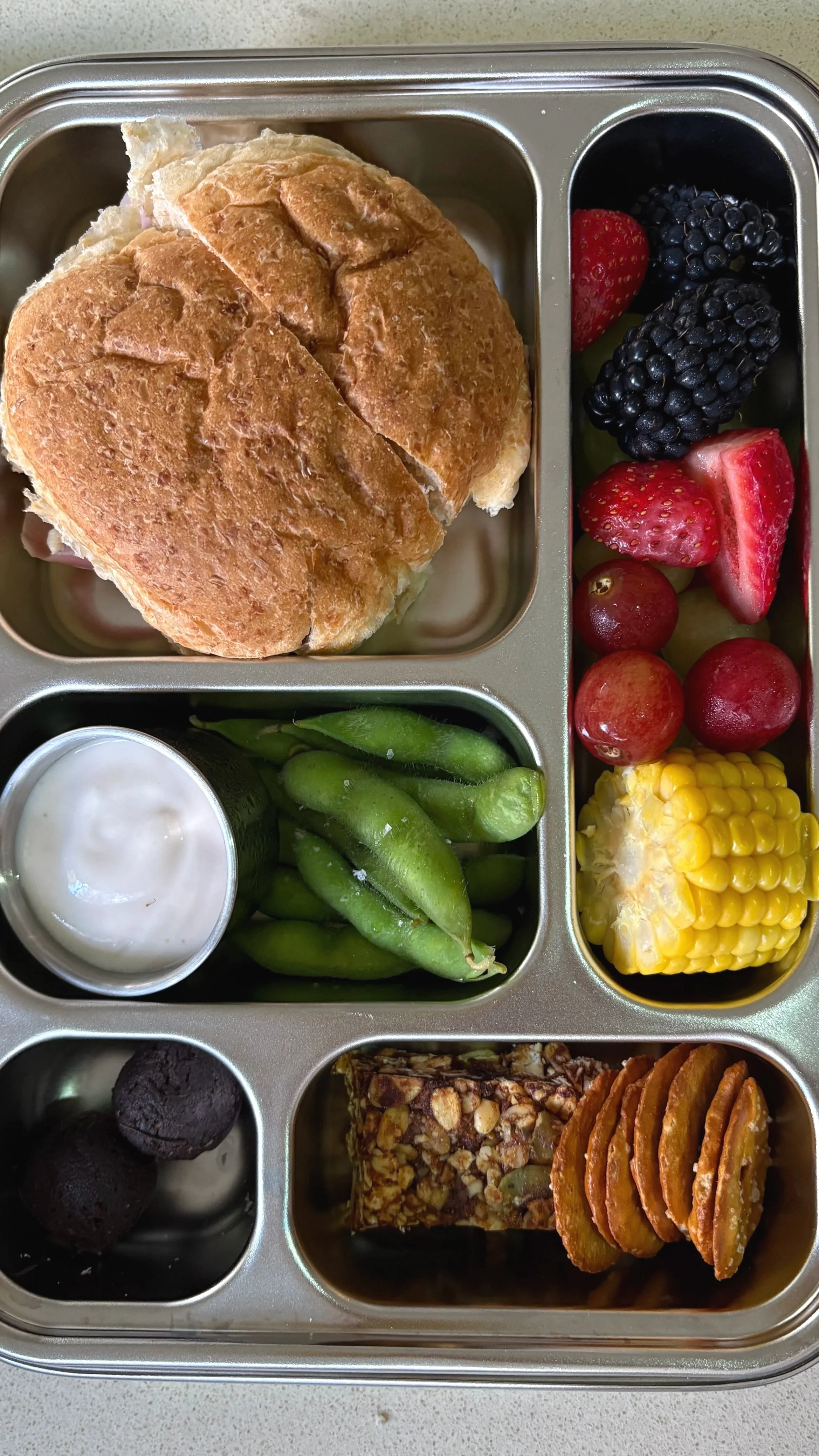 A healthy, easy and balanced low-tox lunchbox with a sandwich, mixed berries, green beans with dip, corn on the cob, chocolate cookies, granola bar, and apple slices.