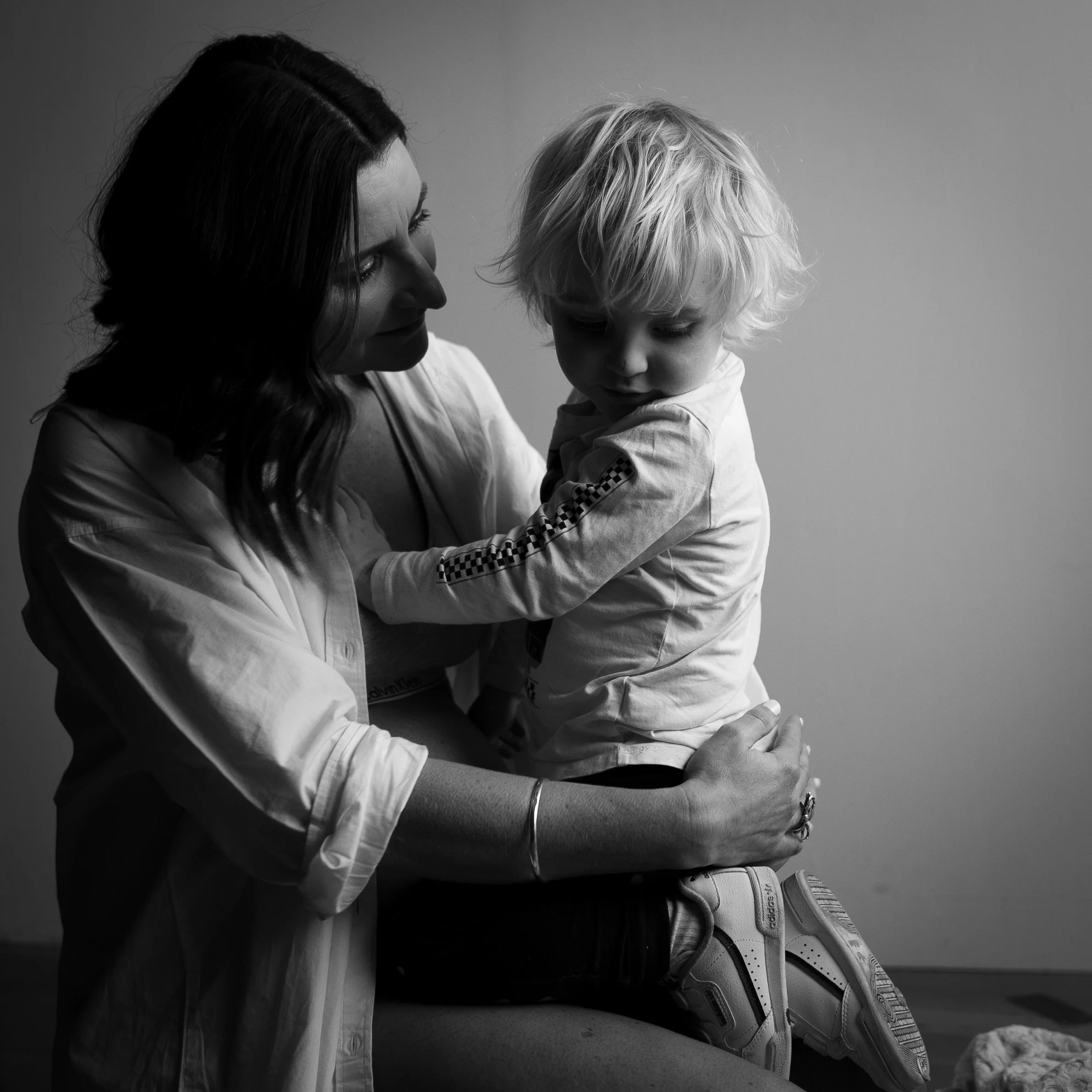Cushla and her young toddler son share a tender moment, with the woman holding the boy on her lap against a plain background.