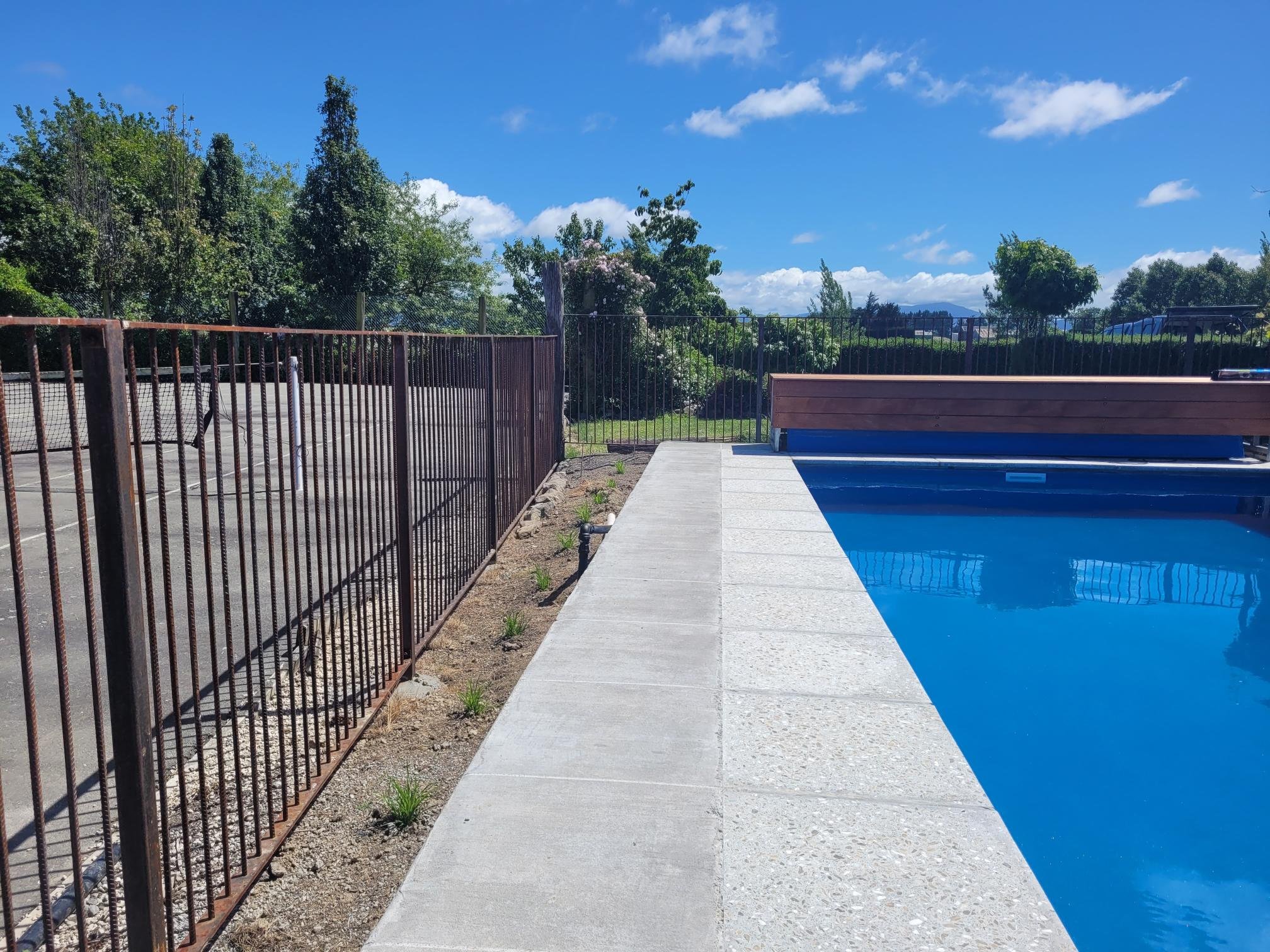 View of a fenced swimming pool with a concrete deck and clear blue water, adjacent to a tennis court, surrounded by green trees and a bright blue sky.