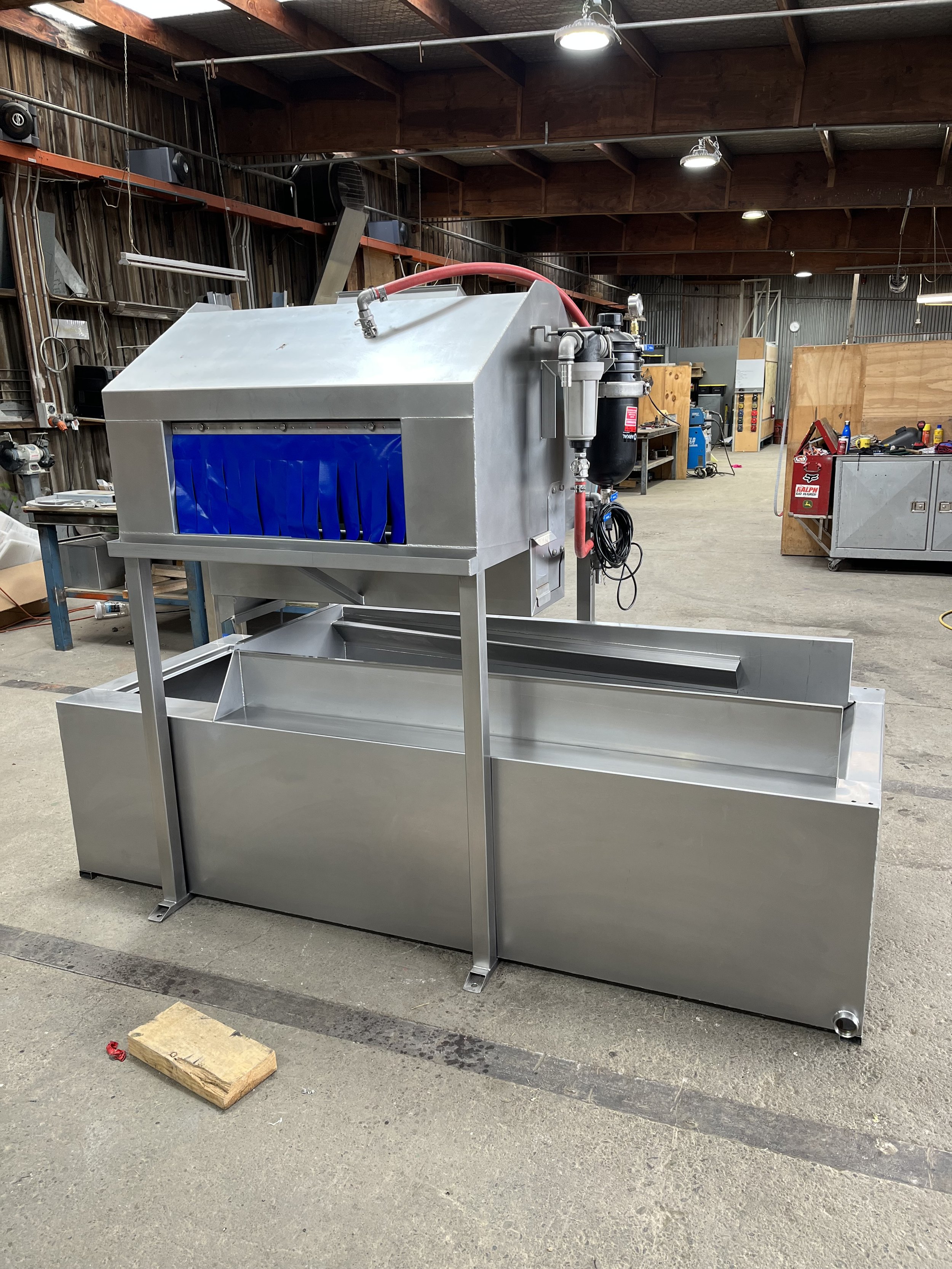Industrial machine in a workshop, featuring stainless steel construction with attached pipes and a blue curtain at the front.
