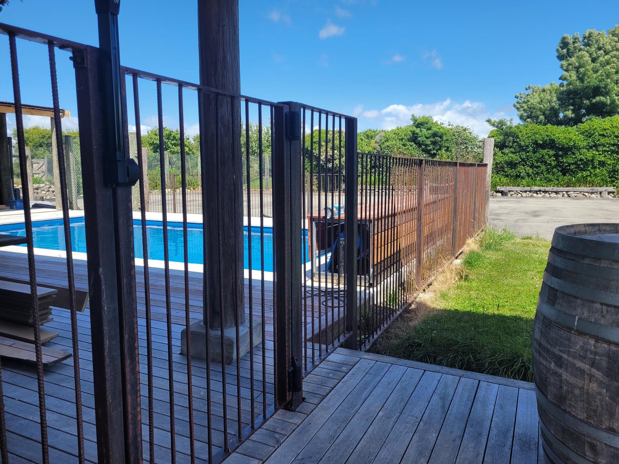 Outdoor swimming pool surrounded by metal fencing and wooden decking, with a blue sky and green trees in the background.