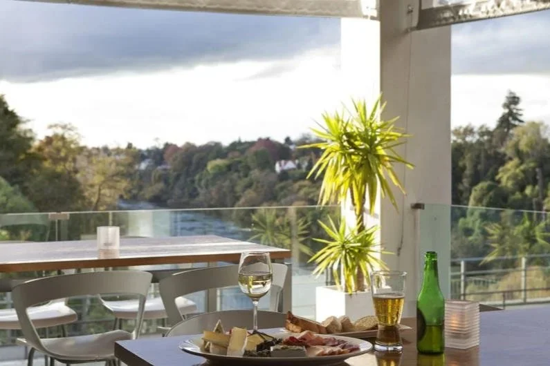 Terraza al aire libre con mesa, sillas, copa de vino, cerveza y plato con vista panor&aacute;mica a los &aacute;rboles y al r&iacute;o al fondo.